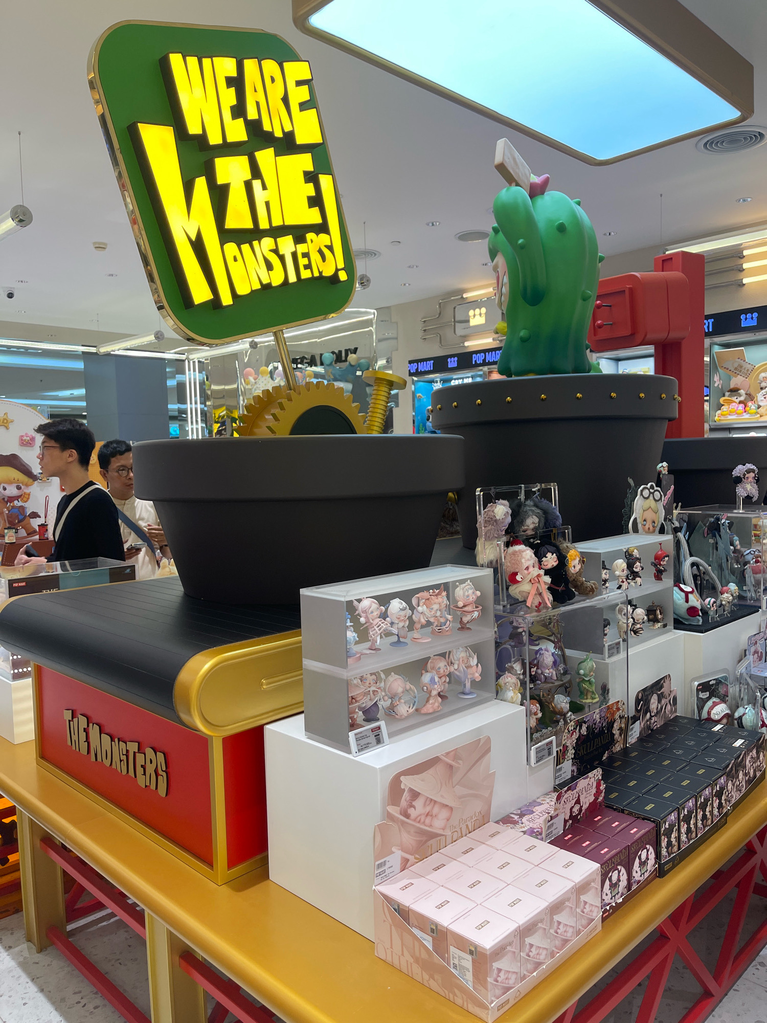 A display of toys and collectibles is set up in a retail environment, likely a store in South Jakarta, Indonesia. A large, illuminated green sign with yellow 3D lettering that reads "WE ARE THE MONSTERS!" hangs prominently above the display. Below the sign, two large black planter-like objects contain a giant green cactus figurine with a pink flower on its head. The display features shelves filled with various collectible figurines, many of which are small, anime-inspired characters. Boxes of these collectible toys are also neatly stacked on the shelves. In the background, two young men are visible, one in a black long-sleeved shirt and the other in a light-colored shirt, appearing to browse the merchandise. The overall environment suggests a modern retail setting with bright overhead lighting and polished flooring.