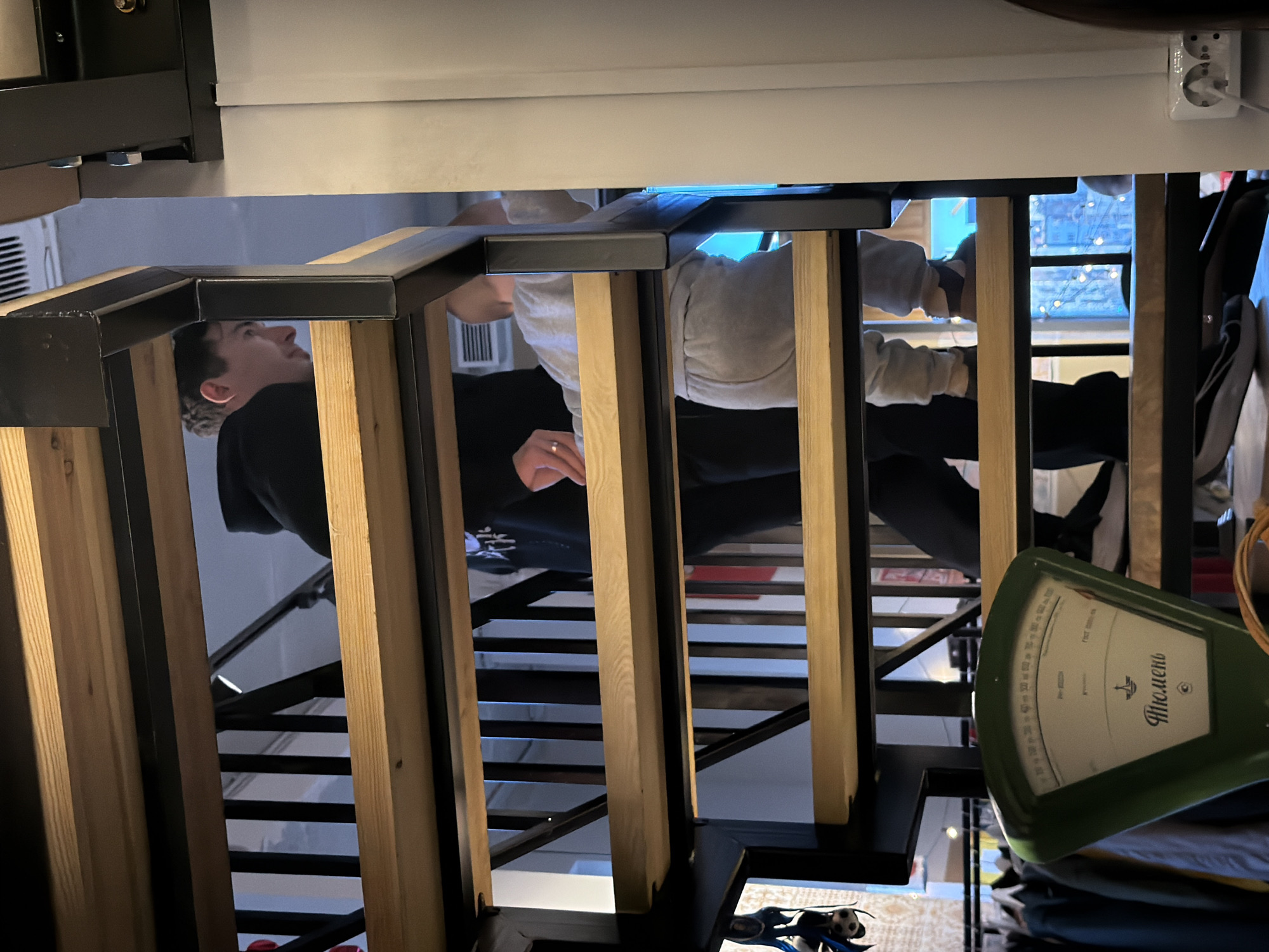 This image captures an indoor scene from a low-angle perspective, looking up towards an elevated platform or loft bed. The structure features a contemporary design with black metal frames supporting light-colored wooden slats or beams, likely forming the underside of the bed or a section of a staircase.
Two individuals are visible through the framework. On the left, a young man with dark hair, wearing a dark hooded top, is seen looking upwards with a neutral expression. On the right, another person is lying down on the elevated platform, dressed in light grey sweatpants and a long-sleeved top. Their left hand, wearing a ring on the ring finger, is visible, suggesting they are relaxing or engaged in an activity on the bed.
Above the structure, a white ceiling or the underside of the platform is visible, featuring an electrical outlet with a white cable plugged in. An air vent is also visible on a wall in the background. Below the main structure, another set of black metal stairs or a ladder can be seen. On the far right of the image, a vintage green mechanical scale with a circular dial is prominent.
The scale displays Cyrillic text, including "Тюмень" (Tyumen) and "ГОСТ 2002-60" (a Soviet-era standard), along with numerical measurements from 0 to 1000, likely indicating weight in grams or kilograms. Below the scale, stacked clothes or fabric items are partially visible. The lighting suggests an indoor setting, possibly in the evening or at night. The scene implies a compact, multi-level living space, consistent with the location context of Izhevsk, Russia.