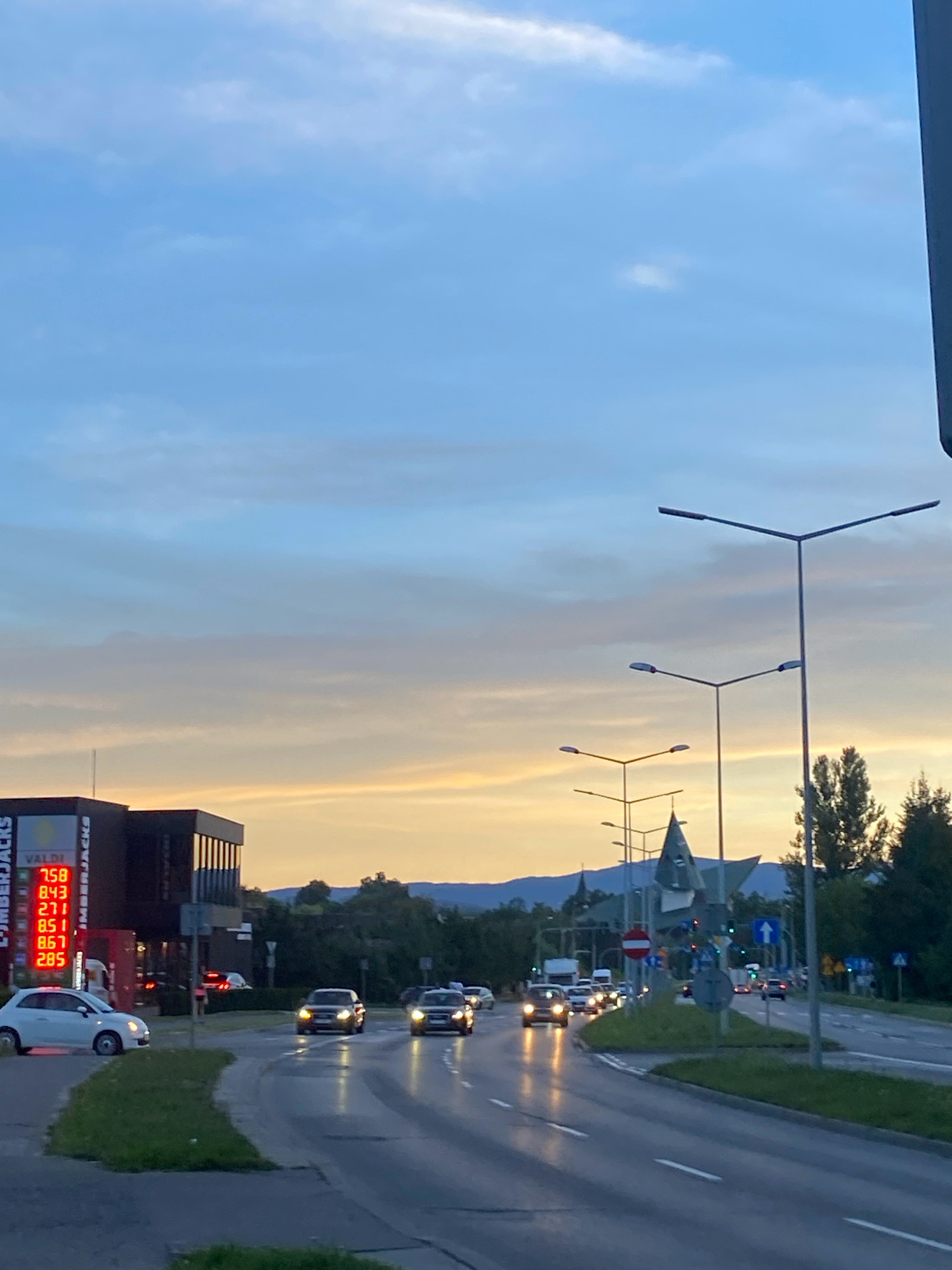 This is a vertical, eye-level, medium-shot photograph taken from the side of a road in Nowy Sącz, Poland, during a transitional time of day, likely dusk or dawn, with soft, diffused natural light. The sky is a pale blue with streaks of orange and yellow near the horizon, indicating the presence of the sun just below the visible frame.
In the foreground, a paved road curves to the right, reflecting the ambient light. Several vehicles are driving on the road, their headlights illuminated. On the left side of the road, a white compact car is parked on a grassy verge. Adjacent to the parked car is a dark, modern-looking building with a large vertical sign that displays the words "TIMBERJACKS" in white letters. Below this, a digital display shows red numbers: "7.58", "8.43", "2.11", "8.67", and "2.85".
Further down the road, on the right side, multiple streetlights with a distinctive Y-shaped design are visible. In the background, there are trees and rolling hills. A church steeple is visible in the distance, beyond the streetlights and the modern building. The overall scene depicts a typical urban or suburban streetscape under a transitional sky. There are no people clearly visible in the image.