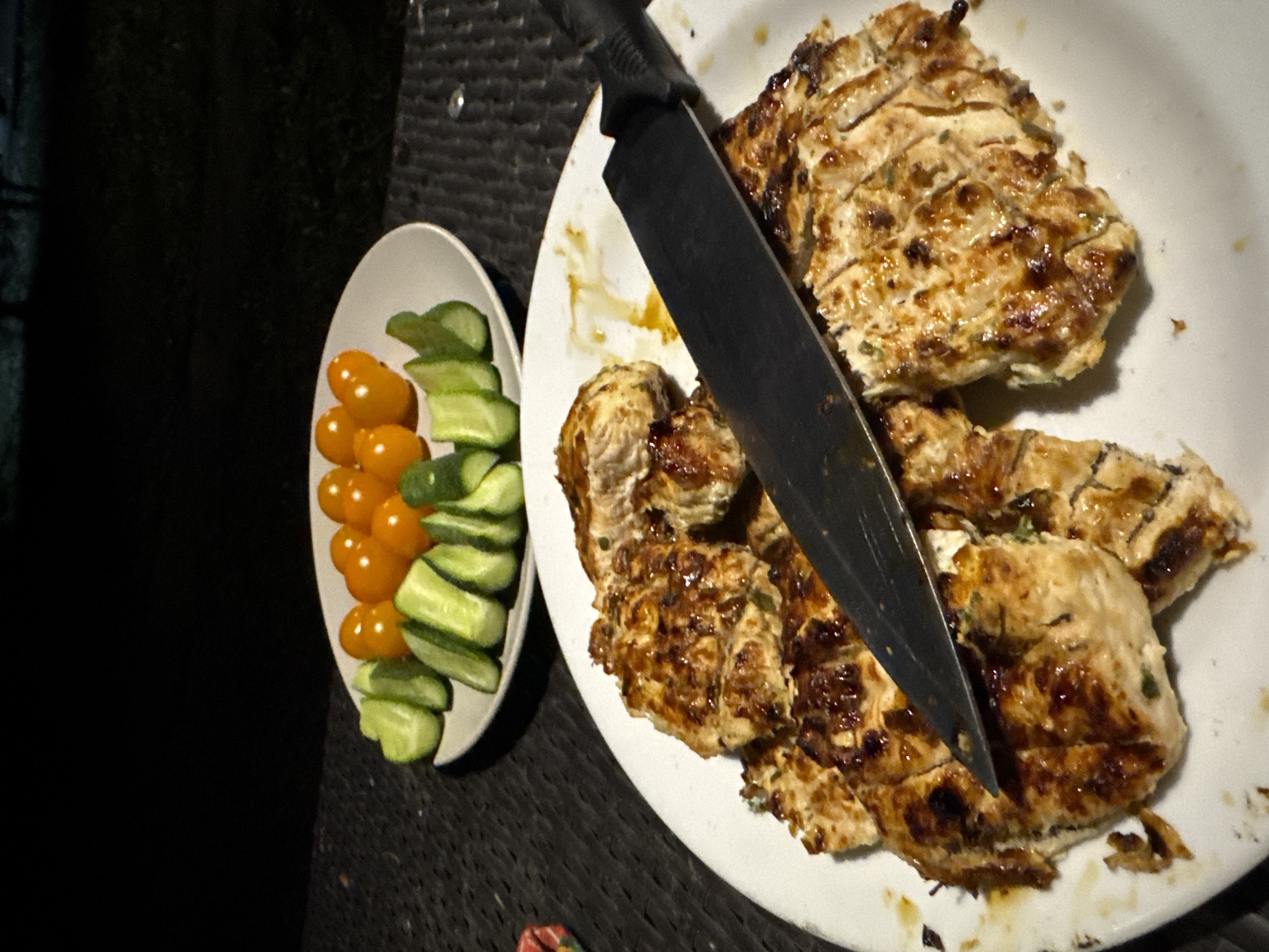 The image presents a prepared meal arranged on a dark, textured surface, possibly a table or outdoor furniture. In the foreground, a large white plate holds several pieces of grilled or pan-fried meat, likely chicken or turkey breast, which has been sliced. The meat exhibits a browned, glossy surface, suggesting a savory marinade or sauce. A large black chef's knife rests on the plate, with its blade extending towards the lower left. To the left of the main plate, a smaller, white oval bowl contains fresh vegetables: a pile of vibrant yellow cherry tomatoes on one side and neatly sliced green cucumbers arranged opposite them. The background is overwhelmingly dark, indicating that the photograph was taken in a low-light environment, likely evening or night, with artificial illumination highlighting the food. No people are visible, and there is no discernible text within the image. The scene suggests a meal ready for serving or consumption, possibly at a casual dinner setting.