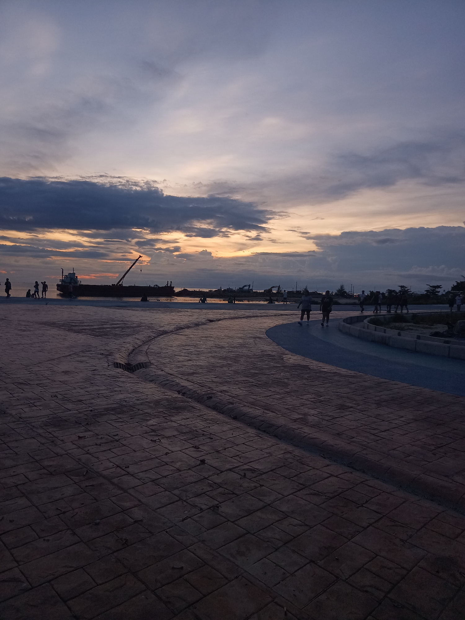 The image depicts a scenic view of a waterfront promenade at dusk. The sky is filled with dramatic clouds, transitioning from a deep blue at the top to hues of orange and yellow near the horizon, suggesting the sun has recently set or is about to rise. Silhouetted figures of people are scattered across the paved area and along the waterfront, some walking, others standing in small groups. A large barge or ship with a crane is visible in the water, anchored near the shore. The foreground is dominated by a textured, dark-colored paved walkway, with a curved blue pathway visible further back. The environment appears to be an open public space, likely designed for recreation and enjoying the coastal scenery. The specific location in Tambon Ko Phangan, Thailand, suggests a tropical island setting, characterized by its maritime elements and potentially a relaxed atmosphere.