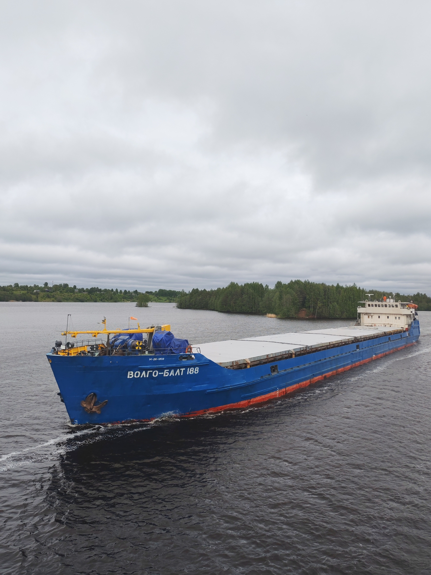 A blue cargo ship with the name "VOLGO-BALT 188" in white lettering is traveling on a river in Devyatiny, Russia under a cloudy sky. The ship is long and narrow, with a flat deck covered in light-colored panels. The hull of the ship is blue with a red waterline. A small cabin structure is visible at the rear of the ship. A wooded shoreline can be seen in the background on both sides of the river. The water is dark and rippled, with white foamy wake trails behind the ship.