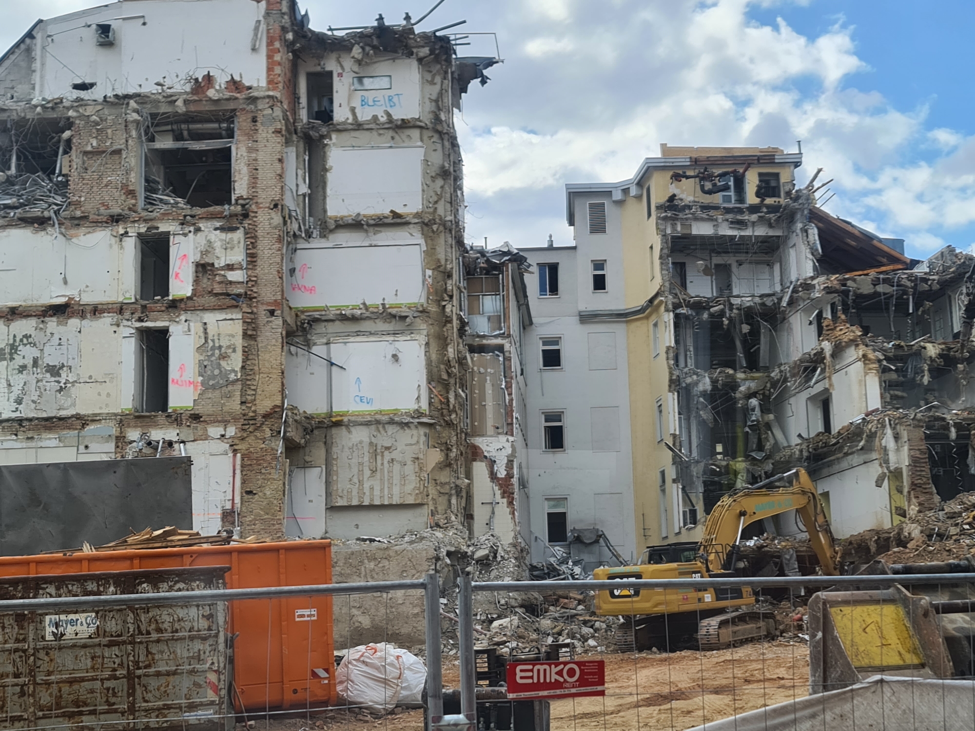 The image captures a demolition site in Vienna, Austria. The main subjects are two partially demolished buildings, one on the left appearing older and made of brick and white plaster, and another on the right that is yellow and white. The demolition is clearly in progress, with debris scattered everywhere and exposed internal structures.
A large yellow excavator is the most prominent object in the scene, positioned within the rubble of the right-hand building, actively engaged in demolition work. In the foreground, two large industrial containers are visible, one orange and one gray, likely used for waste removal. A construction fence separates the viewer from the demolition site.
The weather appears overcast with some patches of blue sky visible, suggesting it is daytime, possibly late morning or afternoon. The overall mood of the image is one of destruction and construction, indicating a transition from an old structure to a new development.
Visible text includes "BLEIBT" and what appears to be "CEVI" in blue spray paint on the left building. On the gray container, "Mayer & Co." is visible. A red sign in the foreground displays "EMKO REITT" with additional text underneath. The scene is a typical urban construction or demolition environment.