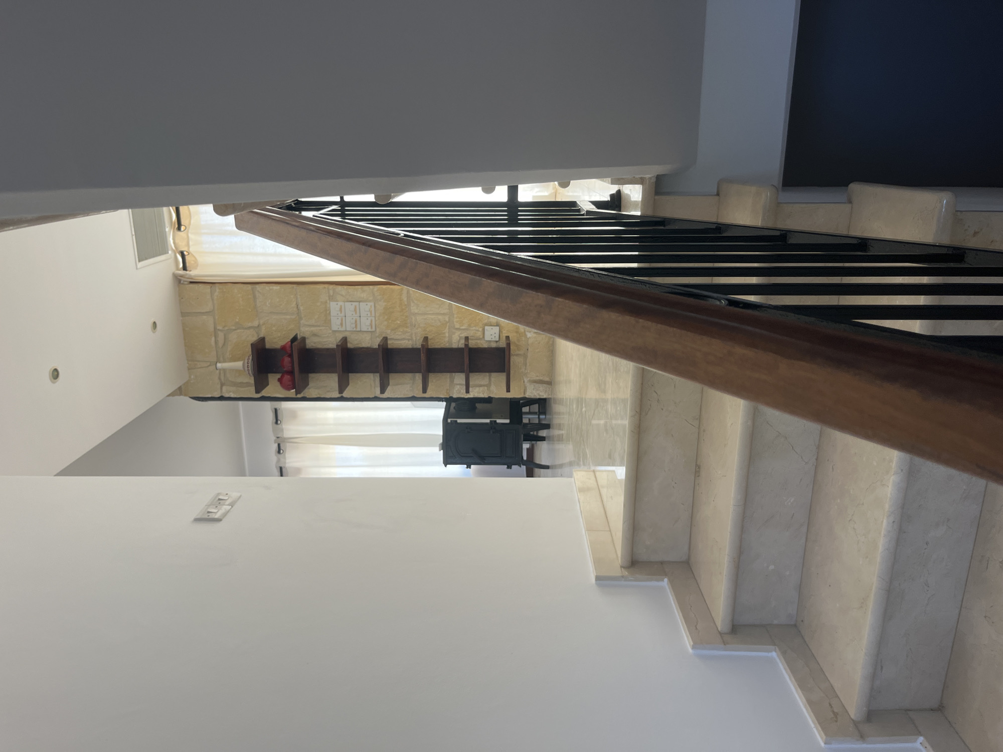 The image depicts a staircase and hallway inside a residence in Aphrodite Hills, Cyprus. The staircase features marble steps with a polished finish and a wooden handrail supported by black metal balusters. The hallway extends into a room with light-colored stone walls and white curtains. A dark-colored wood-burning stove is visible in the background. Electrical outlets are mounted on the stone wall. The overall environment suggests a well-maintained interior of a home.