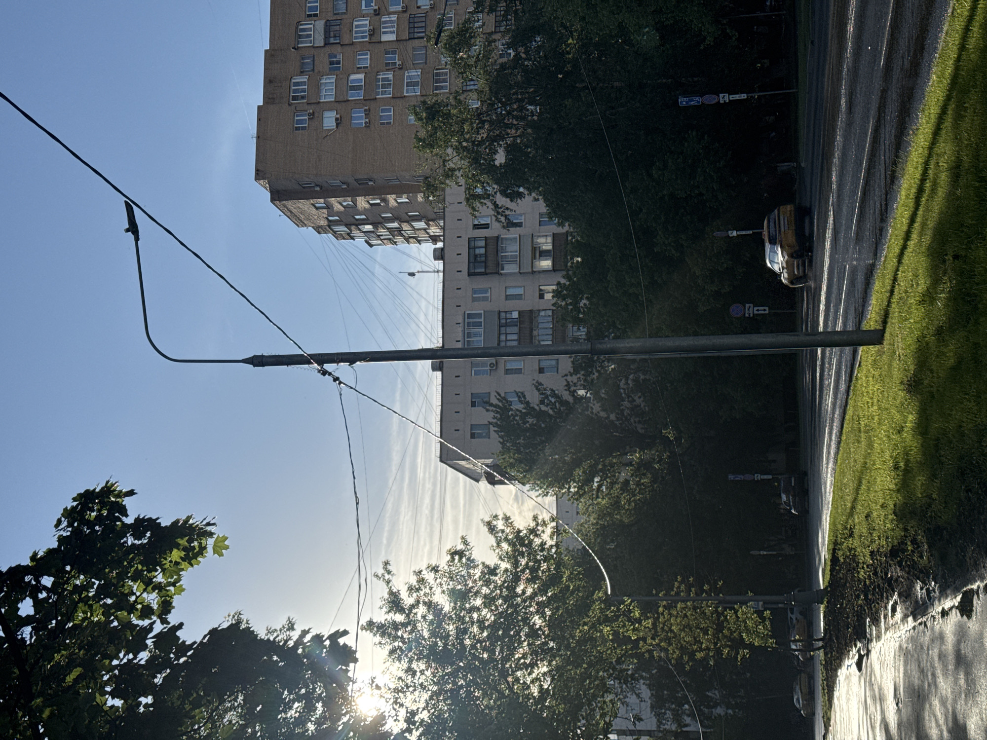 The image shows a street scene in Moscow, Russia. A tall, multi-story apartment building with beige and white facades is visible in the background. Power lines crisscross the sky in the foreground, supported by metal poles. Green trees line the street, partially obscuring the buildings. A street with a curb and sidewalk is visible, with a yellow car parked on the road. The sky is blue with some contrails.