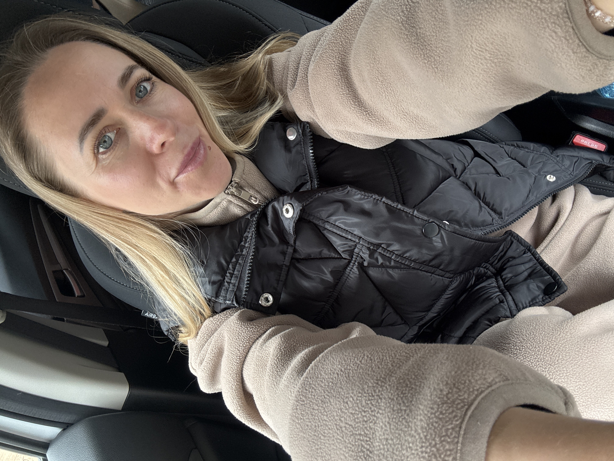 A woman is sitting in what appears to be a car, wearing a beige fleece top and a black quilted vest. The car interior is dark with black seats and trim. The presence of an "AIRBAG" label suggests a safety feature within the car. No information about the location, including Chaikovsky, Russia, can be discerned from this image.