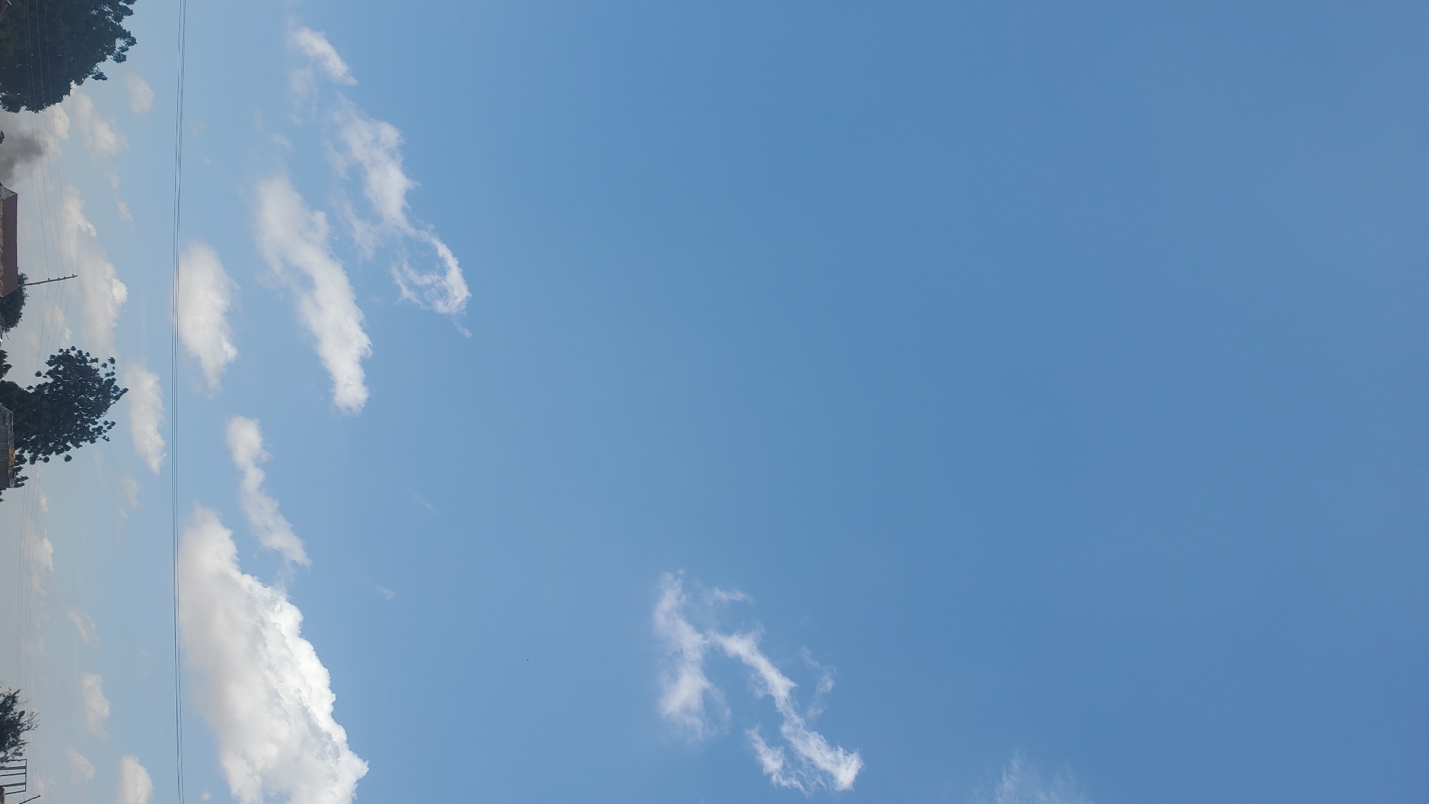 The image shows a clear blue sky with scattered white clouds. In the lower part of the image, there are several trees, power lines, and rooftops, suggesting a semi-urban or rural environment. The scene is likely an outdoor setting in Numan, Nigeria, characterized by open skies and vegetation.