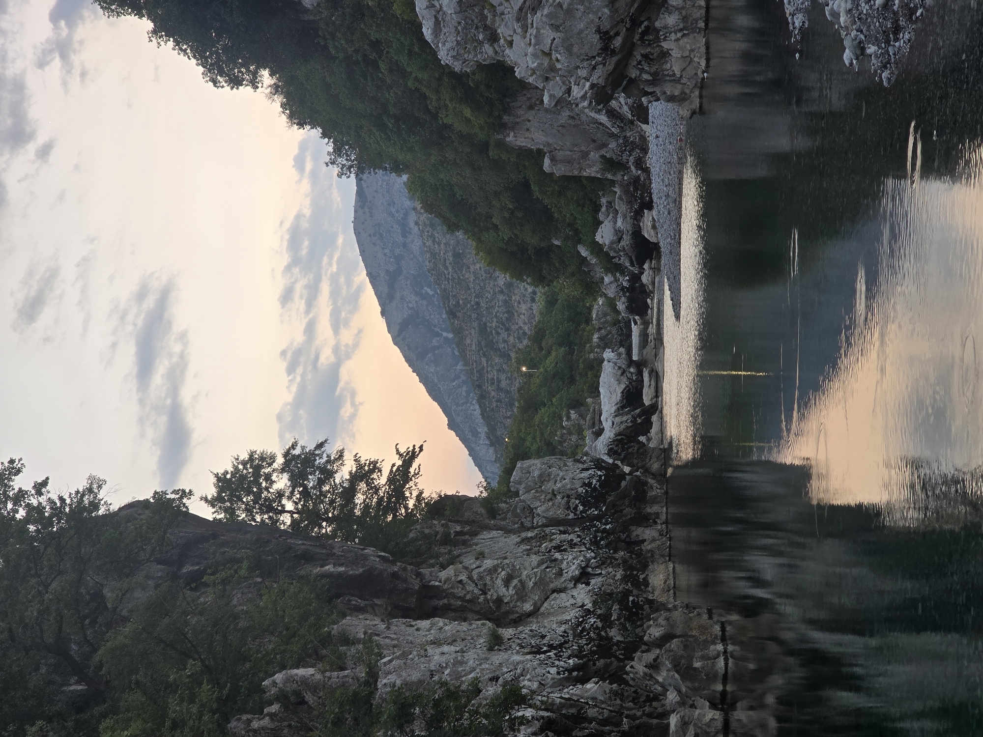 The image depicts a serene river flowing through a canyon at dusk. Towering rocky cliffs covered in lush green trees flank the river on both sides. The water's surface is calm, reflecting the pale sky and the silhouettes of the surrounding landscape. In the distance, mountains are visible, with the sky transitioning through shades of orange and purple as the sun sets. The riverbed is strewn with smooth, grey pebbles. The scene is devoid of any human presence, creating a tranquil and natural environment. This view is characteristic of the natural beauty found in Bioče, Montenegro, where the Zeta River carves its path through dramatic gorges.