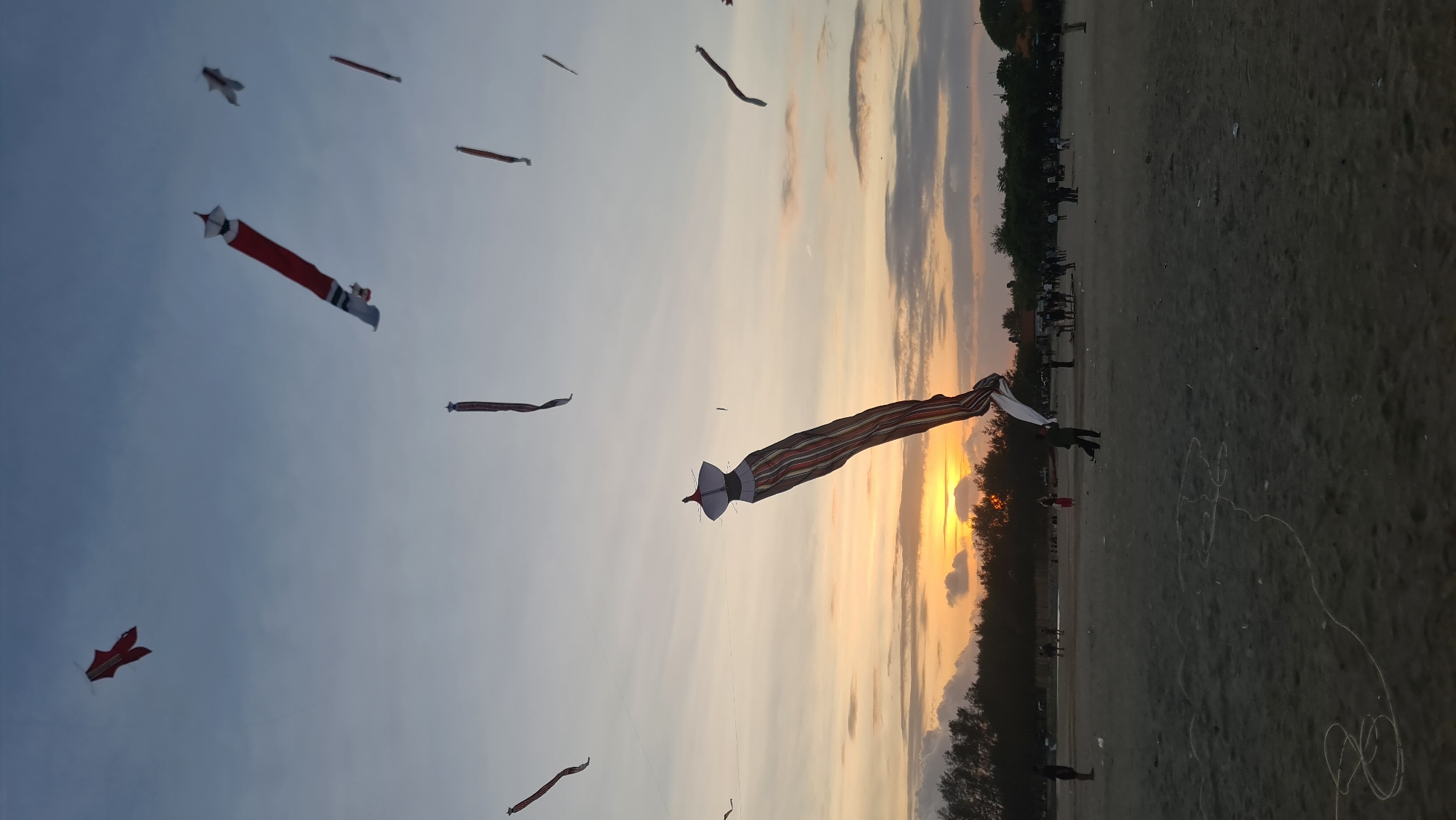 This media file captures a vibrant kite festival or gathering taking place on a beach during sunset. The sky is a dramatic canvas of blues, oranges, and yellows, with wispy clouds indicating a pleasant, perhaps breezy, evening. The setting appears to be a sandy beach in Denpasar, Indonesia, with a treeline and scattered people visible in the background along the horizon.
The main subjects are a collection of large, colorful kites soaring through the sky. One prominent kite, elongated and striped with red and orange, dominates the foreground. It has a distinct, pointed head and a wide tail, suggesting it's a traditional Indonesian kite, possibly a "layangan naga" or dragon kite, though its exact form is unique. Other kites are visible further in the distance, some appearing as long, slender forms, while one smaller red kite has a more bird-like or winged design.
The activity clearly depicted is the flying of these kites. The presence of people, some standing on the beach and others appearing in the distance, suggests a communal event or festival. The overall mood seems to be one of celebration and enjoyment, with the warm glow of the sunset adding a sense of peace and wonder to the scene. The beach itself is sandy and appears to be relatively flat, suitable for kite flying. The time of day is undoubtedly sunset, given the low angle of the sun and the rich, warm colors in the sky. There is no visible text in the media file.