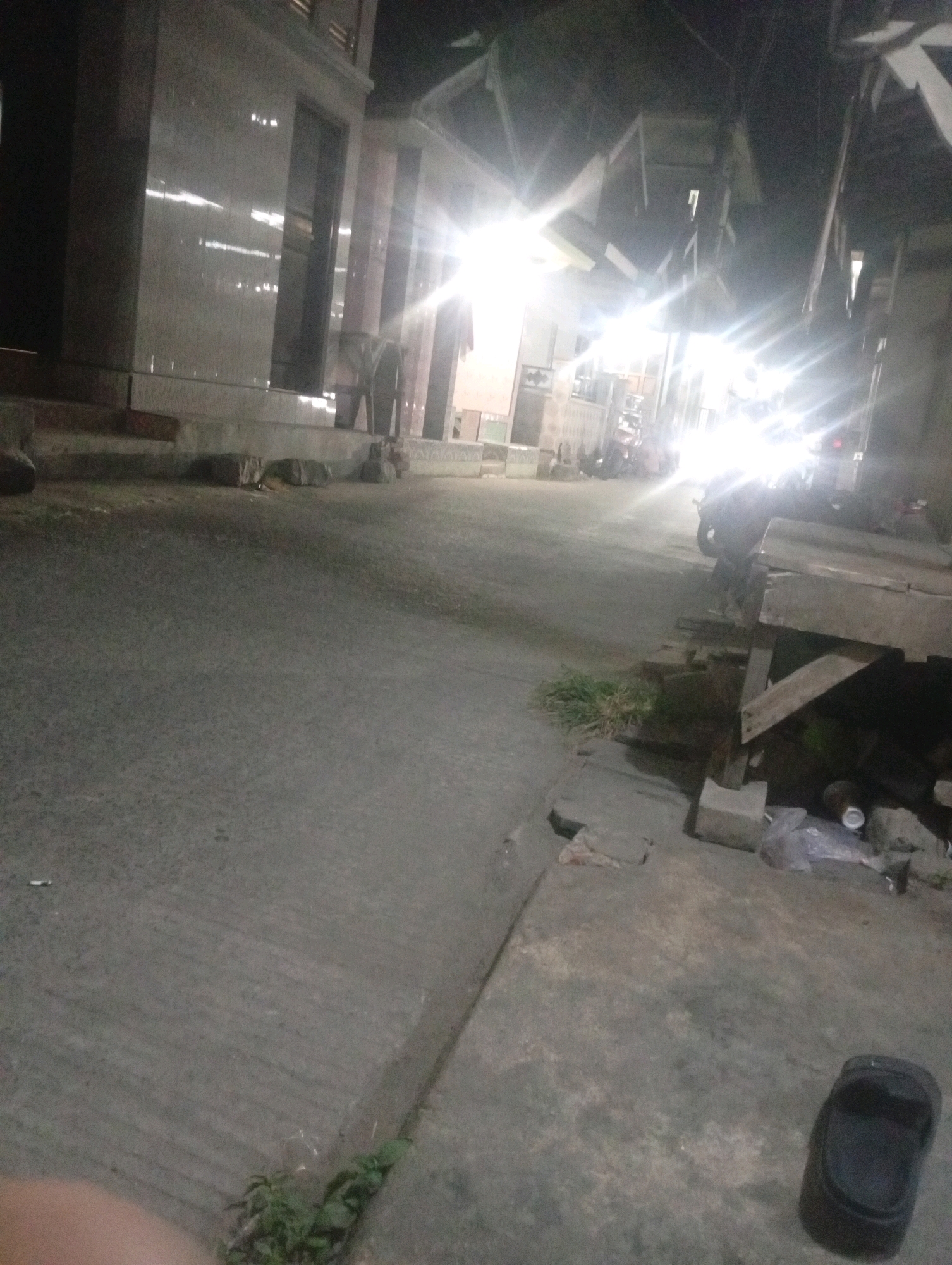 The image depicts an outdoor scene at night, likely in Bajingjowo, Indonesia. A dimly lit concrete pathway runs diagonally across the frame. To the left, a building with reflective tiled walls and arched windows is visible, with a low concrete step leading up to it. Several large, rough-hewn stones are placed along the base of the building. To the right, a wooden structure with a sloped roof is present, appearing somewhat dilapidated. In front of this structure, there are various debris including plastic bags, cans, and some rubble. In the background, bright lights from what appear to be vehicles illuminate the scene, suggesting a street or an area with vehicle traffic. Several motorcycles are parked or moving in the background. A single black sandal lies on the concrete ground in the lower right portion of the image. The overall impression is of a humble, possibly semi-urban or rural environment at night, with signs of everyday life and some disarray.