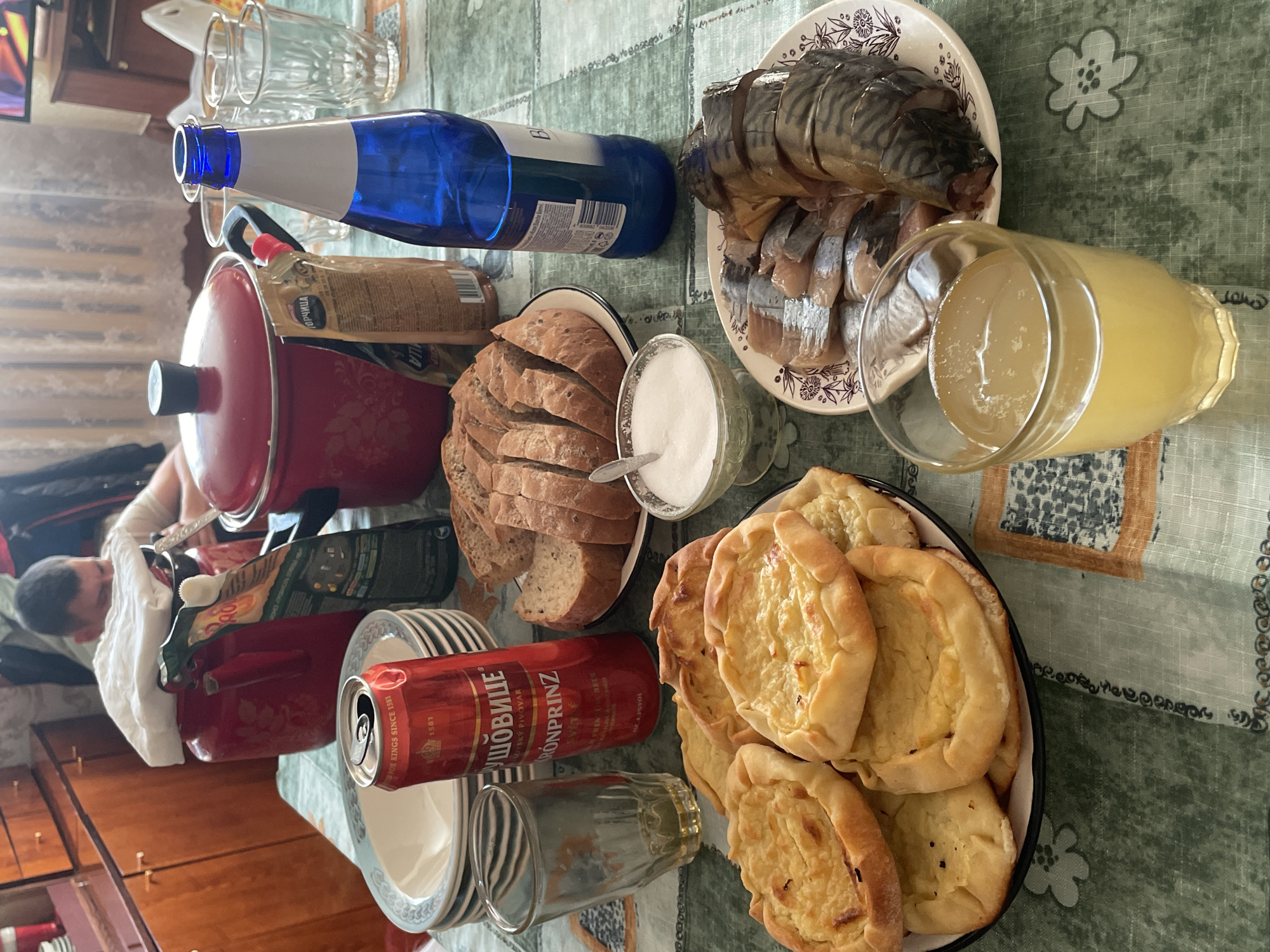 Here's a detailed description of the image:
**Scene:** A table is covered with a green tablecloth. Several food items are arranged on the table. These include sliced bread, a plate of fish (possibly herring and mackerel), a plate of round pastries (possibly filled with cheese), and a bowl of a white substance (likely sugar or salt).
**Objects:**
* A red pot with a black handle
* A bottle of blue liquid
* A can of beer
* Several empty glasses
* Plates stacked near the can of beer
* A glass of a yellow liquid
* A condiment packet.
**People:** The photograph appears to show a person with a white bandage on their arm to the left of the red pot.
**Environment:** The image appears to be taken indoors. There is a wooden cabinet or wall in the background.
**Assumptions:**
* The presence of the items suggests that a meal or gathering is in progress or has just taken place.
* The person on the left is potentially the one who prepared the meal.
**Location:** It is not possible to pinpoint the location as Siva, Russia based only on the image. There are no discernable landmarks or other markers that would indicate the specific city.