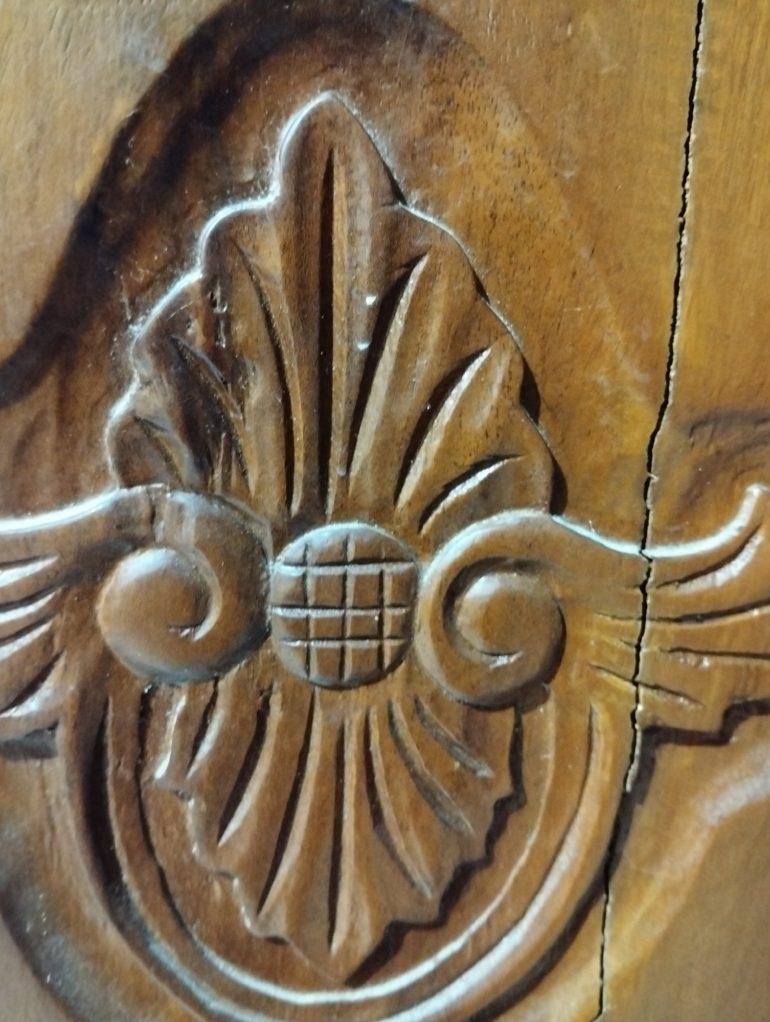 This is a close-up, eye-level, medium shot of a carved wooden detail on a surface, likely furniture or a building facade, in Payagaboh, Indonesia. The carving features intricate, stylized floral or leaf motifs with a central circular element composed of a grid pattern. The wood appears to be dark brown and has a matte finish, with some lighter highlights catching the carved surfaces. A prominent crack runs vertically through the wooden surface on the right side of the image, indicating age or damage. The lighting creates strong shadows that accentuate the depth and texture of the carving. There are no people or other discernible objects in the frame.