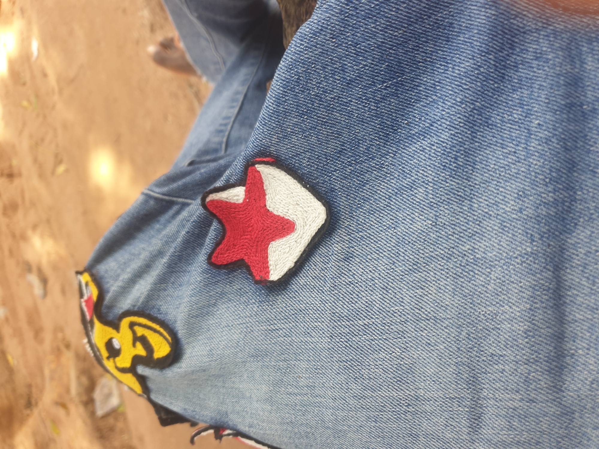 The image shows a close-up of blue denim jeans with embroidered patches. One patch is a stylized yellow and black character with red accents, possibly from a cartoon or anime. Another patch is a red and white star shape. The background is out of focus, appearing to be a sandy, outdoor environment with dappled sunlight. A person's bare foot is visible in the upper right portion of the image. The scene is likely outdoors in a warm climate, such as Jalingo, Nigeria, given the sandy ground and casual attire.