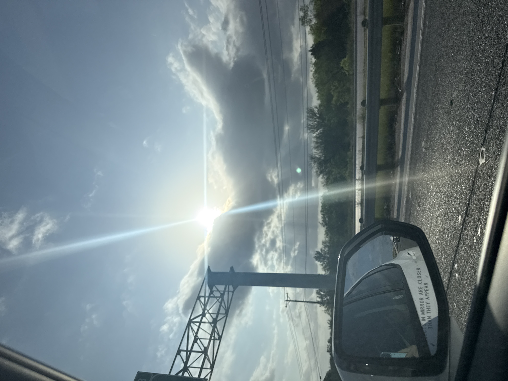 The image depicts a view from inside a vehicle, likely on a highway or major road. The sun is bright in the sky, partially obscured by clouds. A metal tower structure is visible on the left side of the frame. The road surface is wet, and a guardrail runs along the side. The side mirror of the vehicle reflects a white car behind. Based on the presence of a metal tower and highway infrastructure, combined with geographical metadata for the image, it is consistent with the area around New Braunfels, Texas, USA.