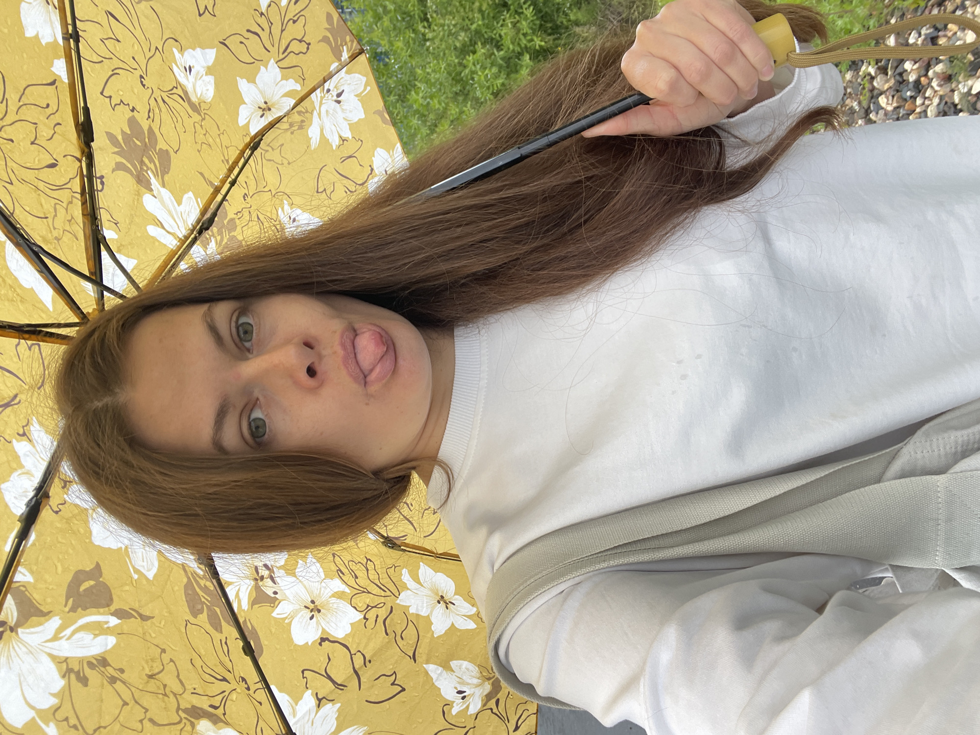 A woman with long, brown hair, is holding a yellow umbrella with a floral pattern, and sticking her tongue out at the camera. She is wearing a white sweatshirt and a light gray bag is slung over her shoulder. The background shows green foliage and a gravel path. The image was taken outdoors, possibly in a park or garden.