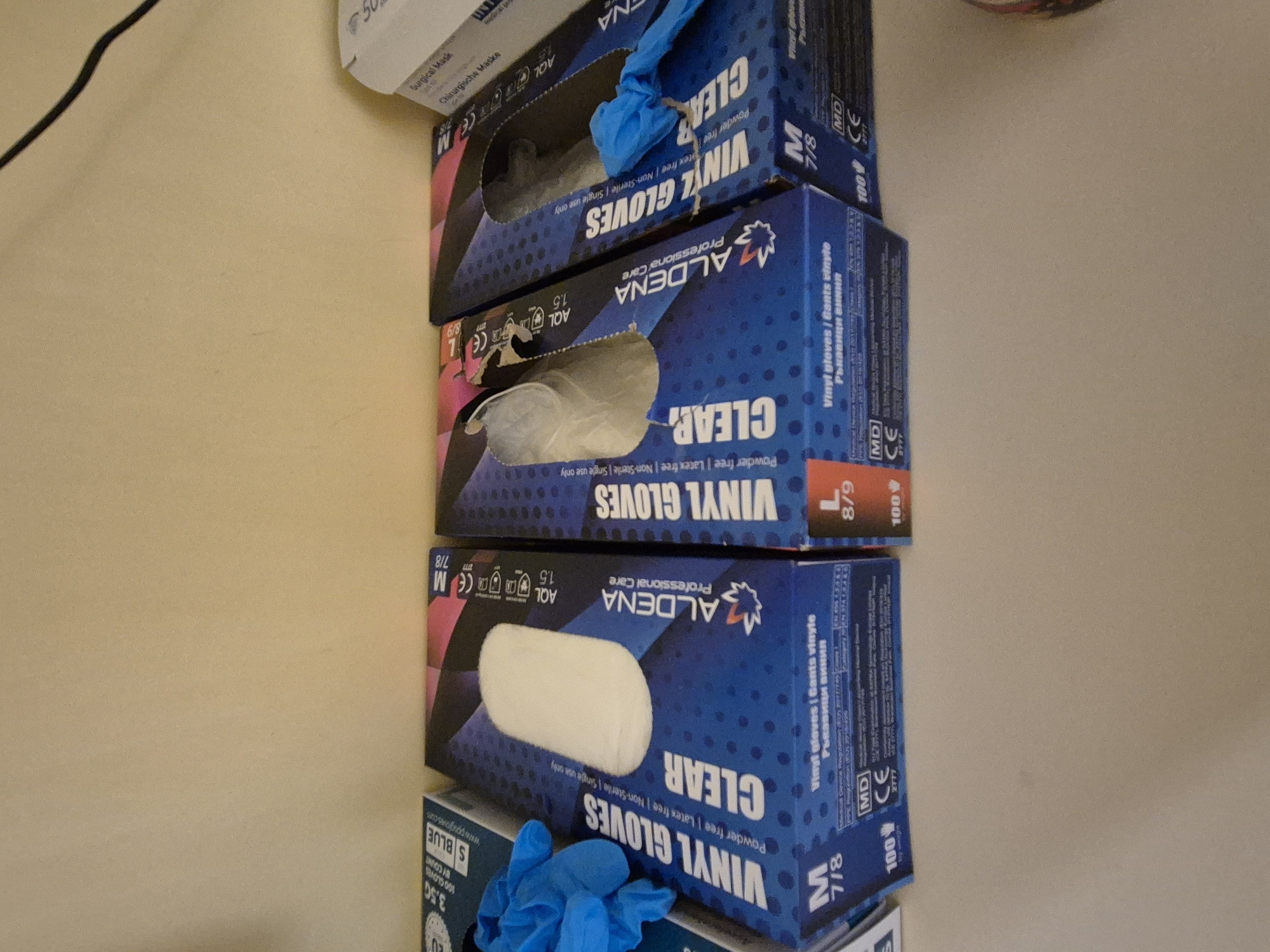 The image displays several boxes of vinyl gloves on a shelf, with a partial view of a surgical mask box to the right. The gloves are of the brand "ALDENA Professional Care" and are clear. Boxes are labeled with sizes such as "M 7/8" and "L 8/9". The environment appears to be indoors, with a plain, light-colored wall in the background. The lighting suggests an indoor setting, possibly a storage area or retail environment. There are no people visible in the image.