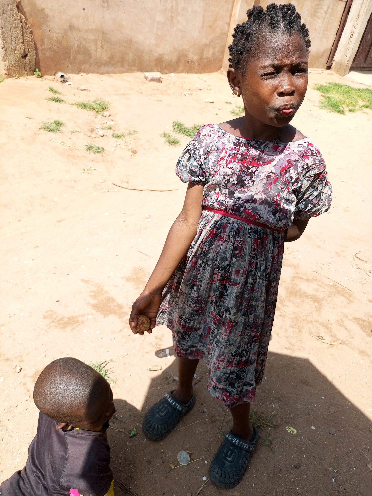 Two young children are standing on dry, sandy ground in front of a rough, light-brown wall. The child on the right, a girl, is facing the camera and is looking to the side with her mouth puckered and one eye squinted. She has braided hair and is wearing a short-sleeved, patterned dress with red, black, and gray designs, cinched at the waist with a thin red belt. She is holding a small, round, brown object in her right hand. The child on the left, who appears to be a boy, is facing away from the camera and is looking down. He has a shaved head and is wearing a dark-colored t-shirt with yellow and pink accents. Both children are wearing dark, slip-on shoes. Sparse, dry vegetation is scattered on the ground. The environment appears to be an outdoor, possibly rural or underdeveloped, area in Tempure, Nigeria, characterized by the sandy terrain and rudimentary wall.