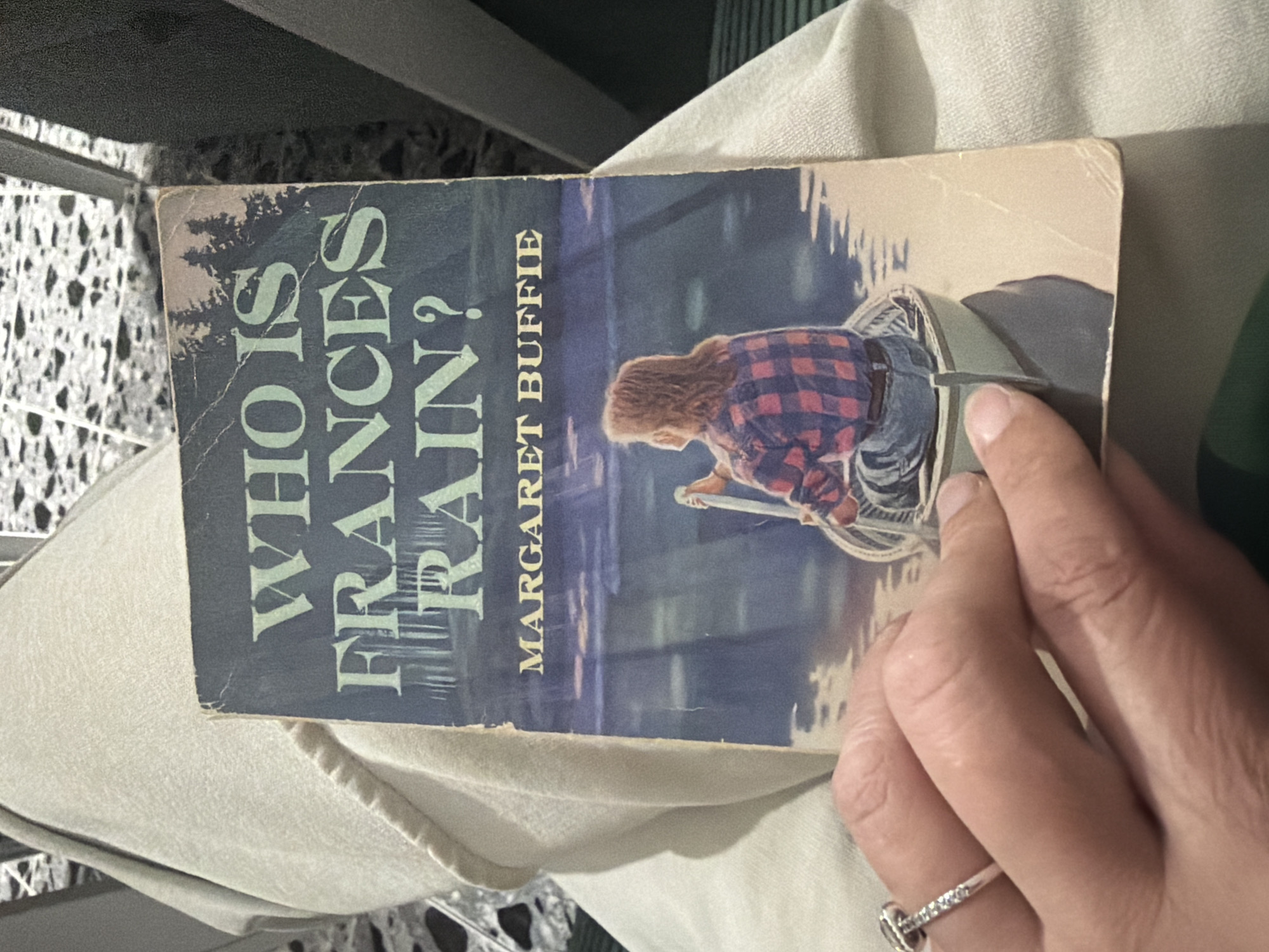 A book titled "Who is Frances Rain?" by Margaret Buffie is partially held open by a hand wearing a diamond ring. The book is set against a background of a person's lap covered in light-colored clothing, with a glimpse of tiled flooring nearby. There is no indication of this image being in Tbilisi, Georgia.