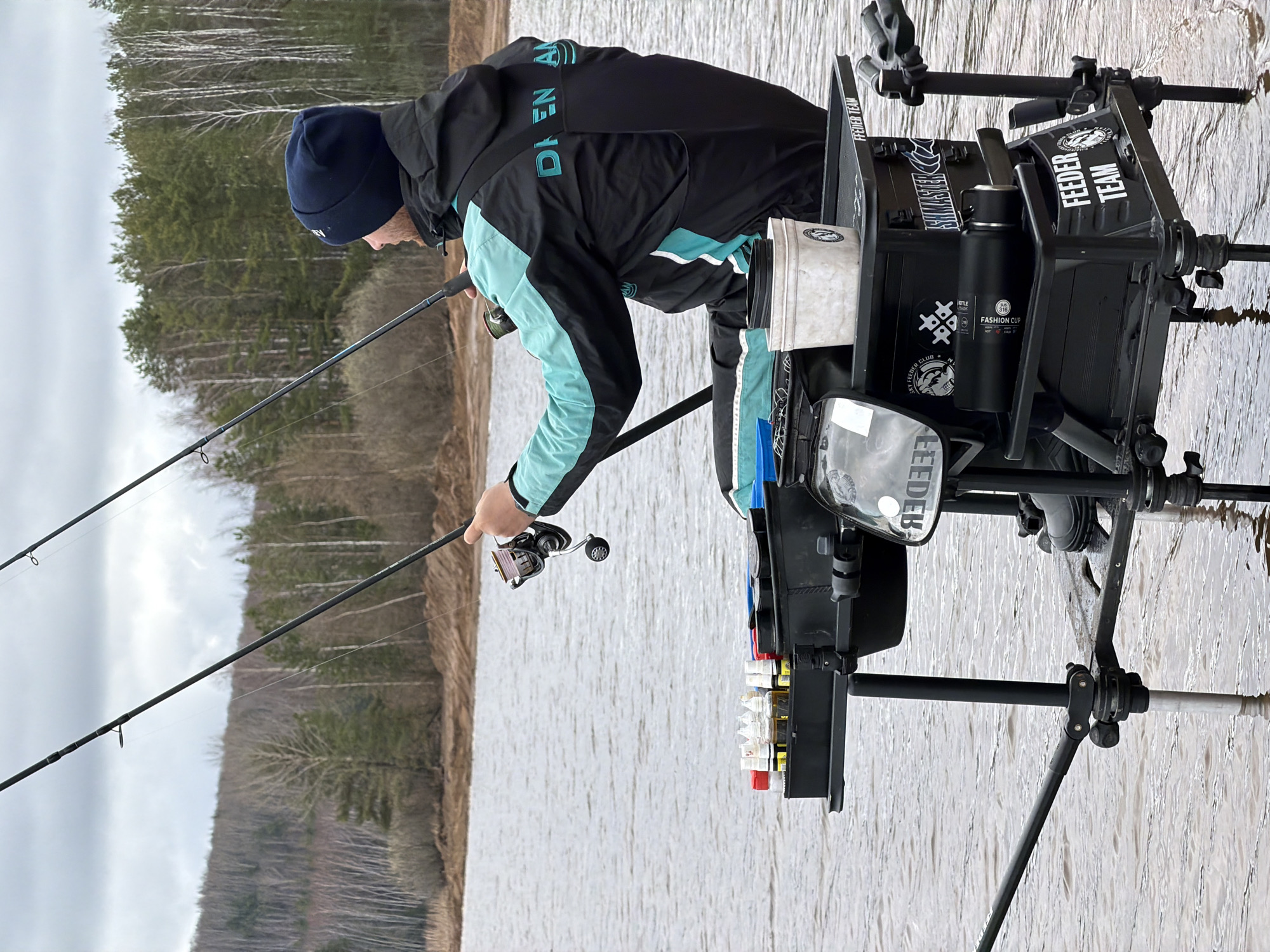 A man is fishing on the shore of a body of water. He is wearing a dark blue beanie, a black jacket with light blue accents, and black pants. He is operating a fishing rod and reel. Behind him is a complex setup of equipment, including a seat, storage containers, and a platform with supporting legs that appear to be submerged in water. The scene is on the edge of a body of water, possibly a lake or river, with trees in the background. The location is Chaikovsky, Russia.