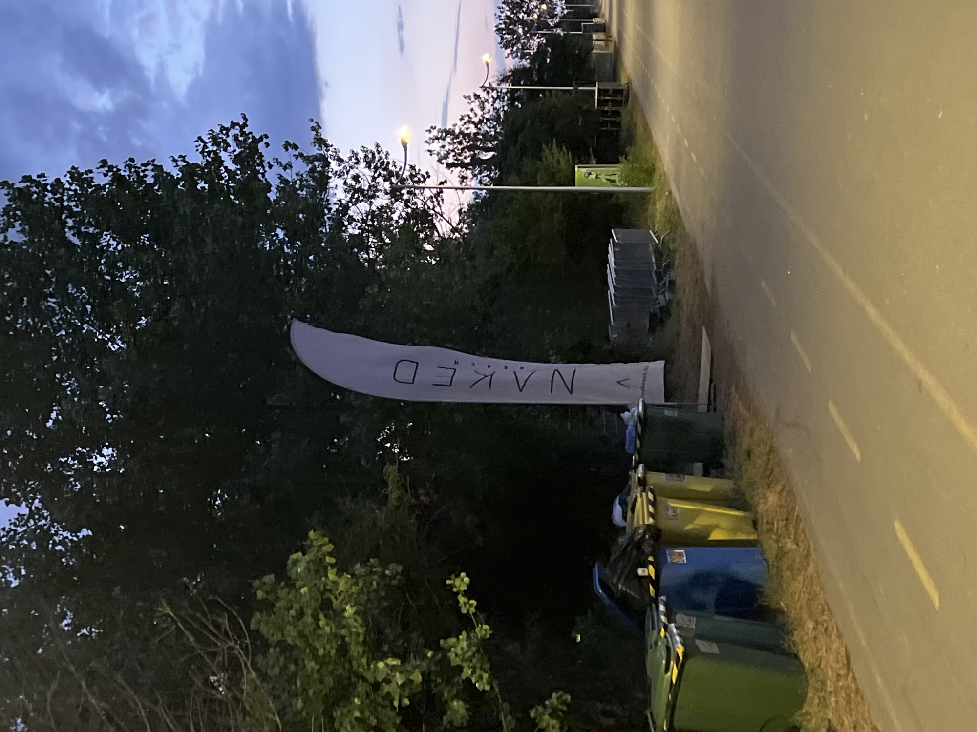 An eye-level, medium shot captures a scene at dusk on an empty, paved pathway lined with streetlights, in an outdoor urban environment. On the left, a row of colorful trash bins, including green, blue, and yellow, are situated on a grassy patch. Behind the bins, a tall, white flag with "NAKED" printed vertically in black lettering stands prominently, a few stacked silver barricades are visible behind it. Dense trees with dark foliage form a backdrop to the left and center of the image, while on the right, the pathway recedes into the distance, illuminated by intermittent streetlights and bordered by more trees. The sky above is a gradient of dusky blues and purples, with scattered clouds. The overall impression is one of an organized, yet deserted, outdoor venue as day transitions into night.