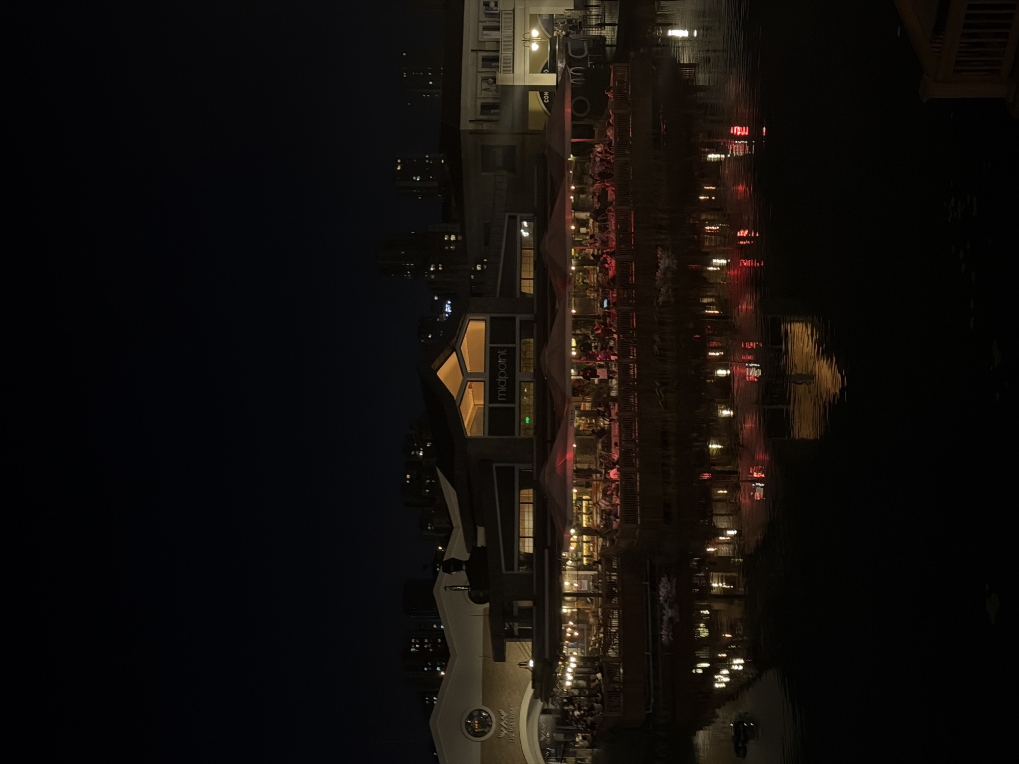 The image captures a night view in Küçükçekmece, Turkey, with a waterfront scene dominated by the reflection of buildings and lights on the water. A multi-story building with a sign that reads "midpoint" is illuminated and surrounded by outdoor dining areas with visible patrons. The reflection of red and yellow lights, likely from restaurants or street lamps, shimmers across the water. Other buildings and city lights are visible in the background.