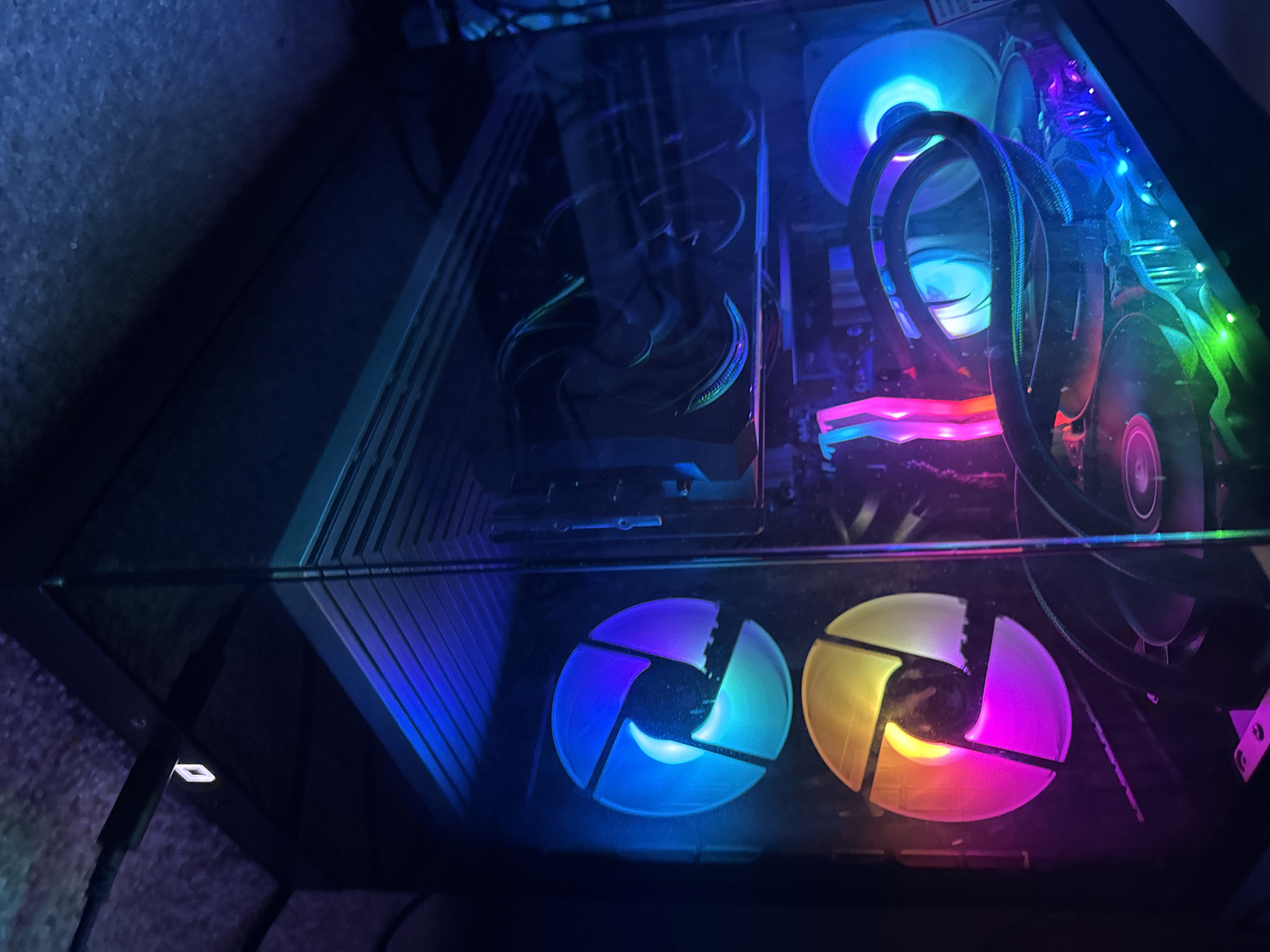 The image shows the interior of a desktop computer, likely located in Glendale Heights, United States of America. The components visible include multiple RGB fans with various color patterns (blue, yellow, pink), a liquid cooler with tubes, and a graphics card. The computer case has a glass or acrylic side panel, allowing a view of the illuminated internal components. The overall environment appears dimly lit with cool color tones.