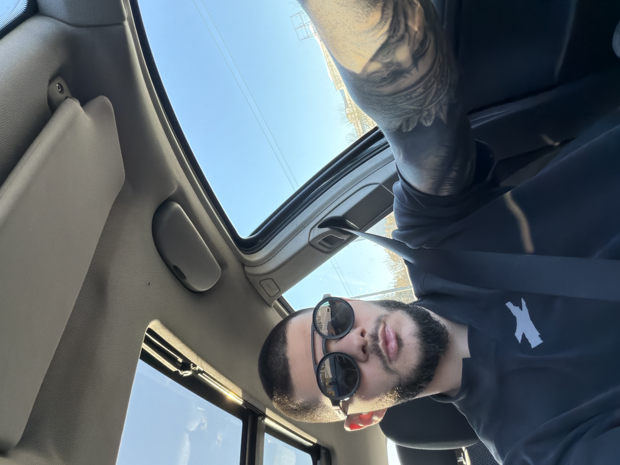 A low-angle, selfie-style photograph shows a man wearing sunglasses in a car with an open sunroof. The sky is visible through the sunroof and side windows. The interior of the car is gray, with a sun visor partially visible. The man has a dark beard and a shaved head. His left arm, featuring a tattoo, is positioned above him. He is wearing a black sweatshirt with a white logo on it. The location in the city of Sofia, Bulgaria, cannot be determined from this image.