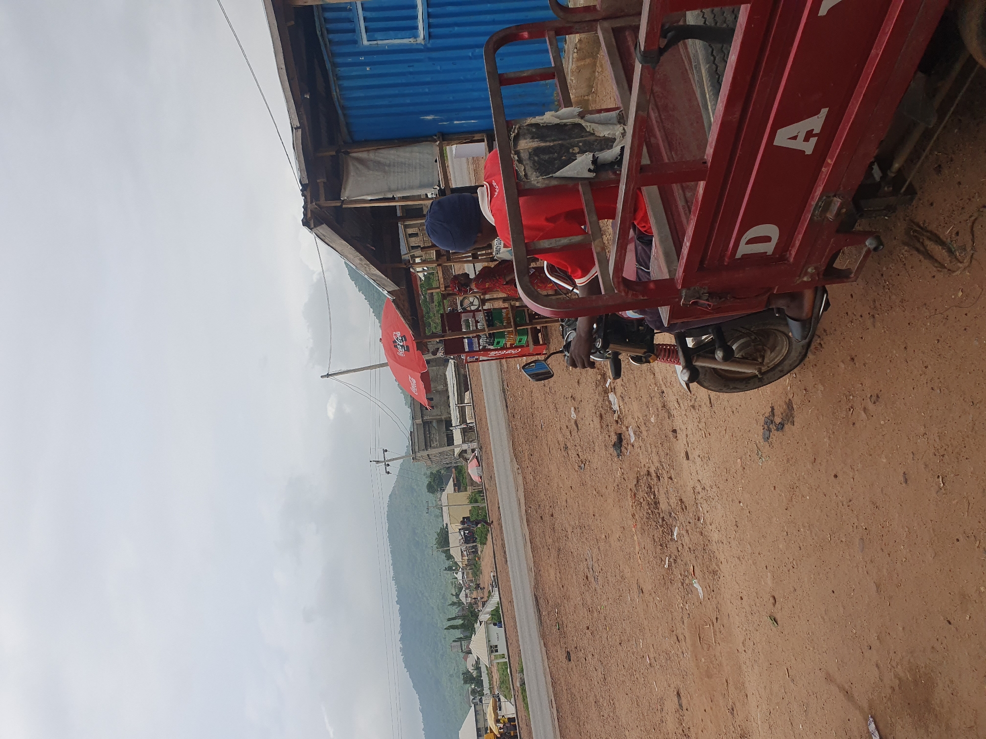 A red, three-wheeled vehicle, often referred to as an "okada" or "keke napep," is parked on a dirt road in Lokoja, Nigeria. The driver, wearing a red shirt and a blue beanie, is seated in the vehicle, facing away from the camera. The vehicle's cargo bed is empty and shows some wear and tear.
Behind the vehicle, a corrugated metal building with a blue facade stands next to a smaller structure with a wooden roof. A red umbrella, possibly a business advertisement, is set up in front of the building. The background reveals a street with a few other vehicles and buildings, and a lush green hill under a cloudy sky. The ground is covered in reddish-brown dirt, with scattered debris like plastic bags and other litter. The overall scene suggests a commercial area or a roadside stop in a developing urban environment.