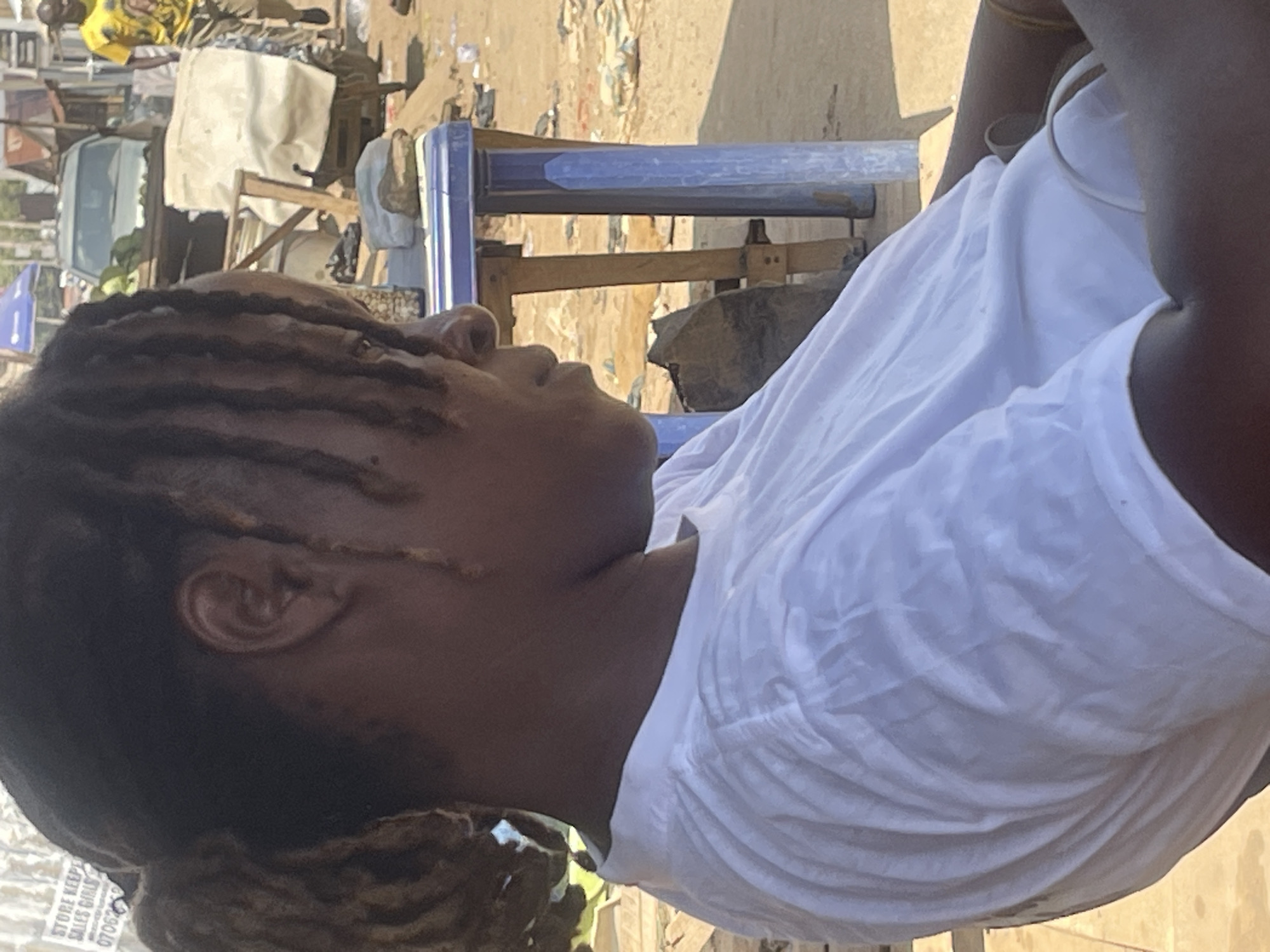 An outdoor market scene in Abuja, Nigeria, features a woman in the foreground wearing a white t-shirt, her dreadlocks pulled back. She is seated, looking to her right. In the background, a man in a yellow t-shirt walks away from the camera, and a light-colored SUV is parked. The environment appears to be a dusty, open area with market stalls and debris scattered around.
