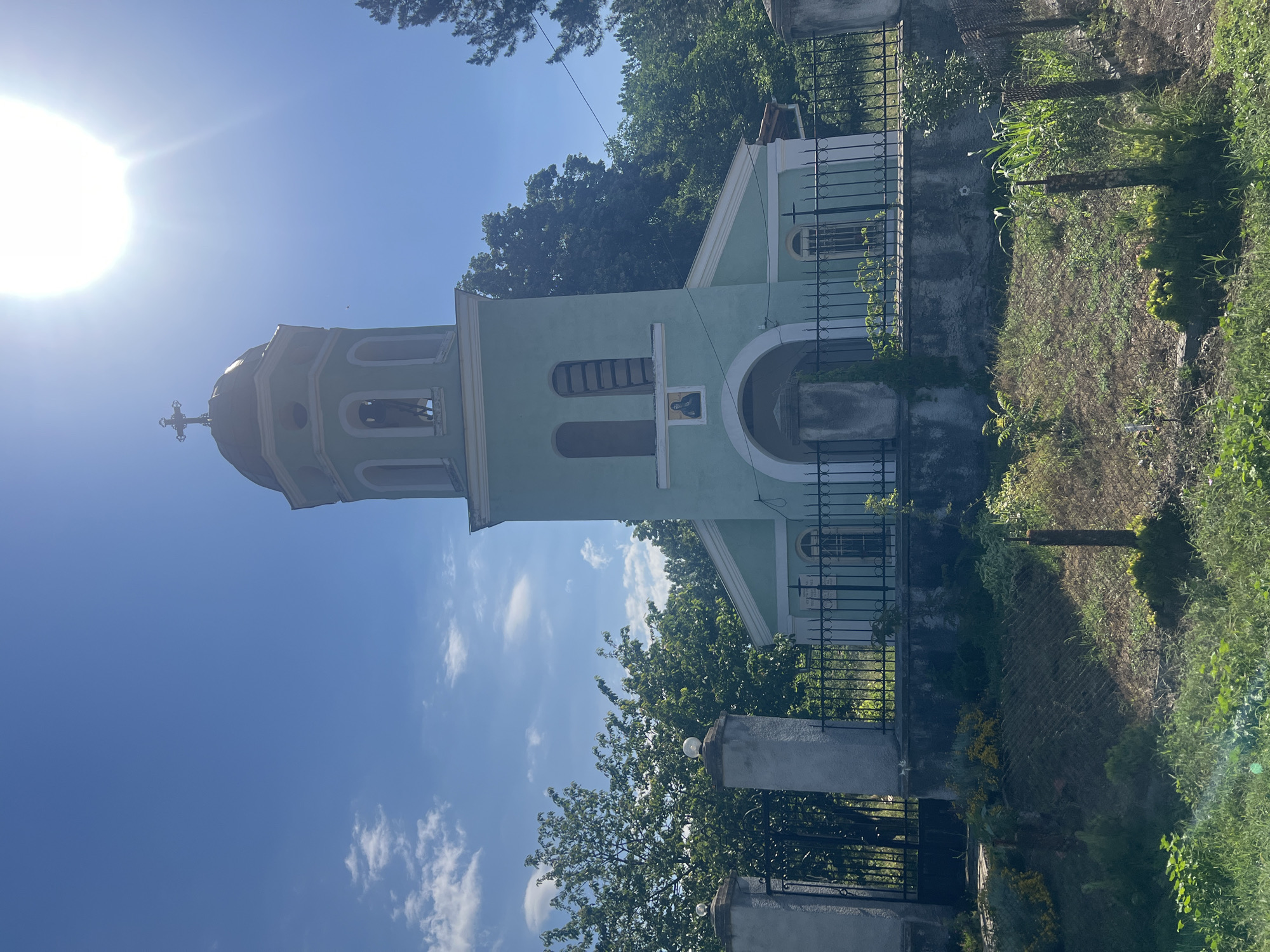 The image shows a light green Orthodox church with a tall, slender bell tower topped with a cross. The church has arched windows and a semicircular entrance. In front of the church is a black metal fence and a low stone wall. The grounds in front of the wall are covered with grass and some wild vegetation. The sky is a bright blue with a few wispy clouds and a strong sun glare in the upper right corner. The overall scene suggests a sunny day in a rural or semi-rural area, likely Zelenikovo, Bulgaria, given the architectural style and prevalence of Orthodox churches in the region. There are no people visible in the image. The environment is dominated by the church structure and lush green trees in the background.