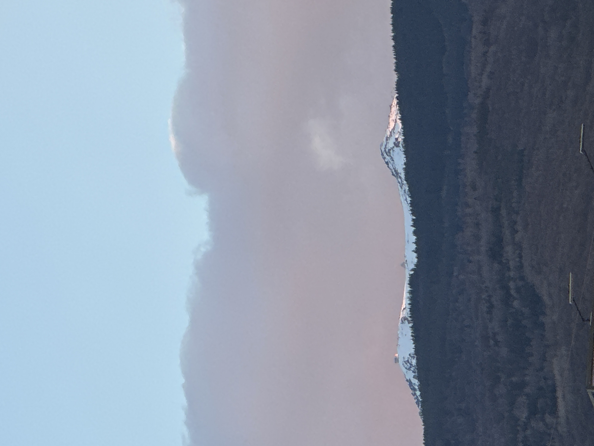 The image displays a serene mountain landscape under a soft, hazy sky. In the upper left, the sky is a clear light blue, gradually transitioning to a paler, diffused tone and light cloud formations or atmospheric haze across the rest of the upper half of the frame. A prominent ridgeline of mountains, heavily forested with dark evergreen trees, extends horizontally across the middle of the image. Patches of snow cap the higher elevations of these mountains, catching a warm, golden-pink light, which suggests either an early morning or late afternoon/sunset glow. The dense tree cover dominates the lower half of the scene. In the far lower right corner, small, indistinct vertical elements, possibly utility poles or distant structures, are faintly visible against the dark treeline. The overall atmosphere is tranquil and expansive, depicting a natural mountain environment with snow-dusted peaks, likely Vitosha mountain near Sofia, Bulgaria. No specific activity or visible text is present.