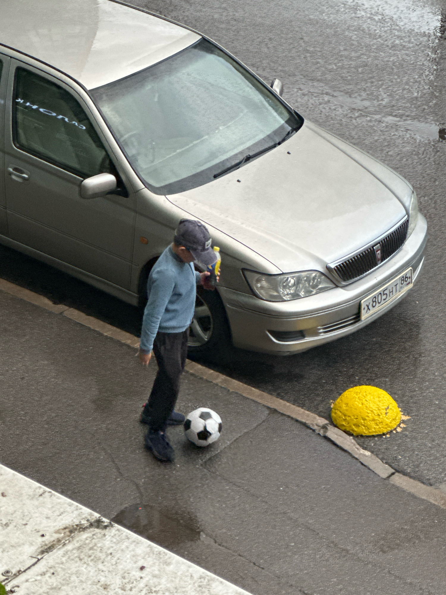 A young person wearing a blue sweater, dark pants, and a dark baseball cap is standing on a wet asphalt street, holding a bottle in their right hand and looking down at a black and white soccer ball at their feet. The person is positioned to the left of a silver sedan. The car has its headlights on and a license plate with the characters "X 805HT 86" visible. A bright yellow, spherical object is on the street to the right of the car. The street appears to be wet, possibly from rain, with puddles visible. A concrete curb and sidewalk are visible in the lower left corner of the image. The scene appears to be in an urban environment.