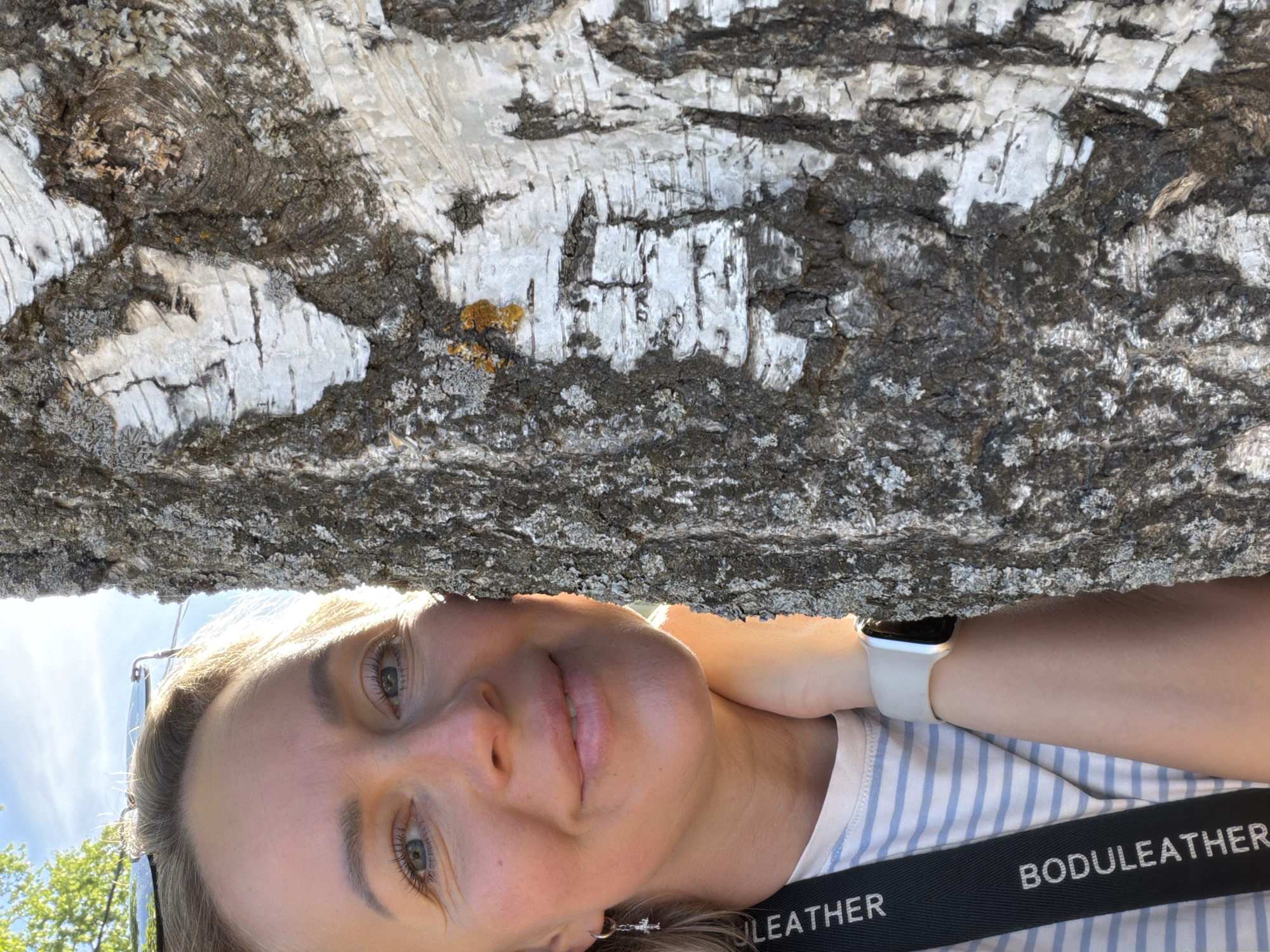 A selfie taken outdoors features a person looking directly at the camera. The subject is beneath a tree trunk, which dominates the upper portion of the frame. The tree trunk is light gray and brown. The person has fair skin, light eyes, and light hair, wearing a striped shirt and a watch with a light-colored band. A black strap with white lettering partially visible reads "BODULEATHER." The setting appears to be in a green area with a blue sky visible in the lower-left corner. The location is Chaikovsky, Russia.