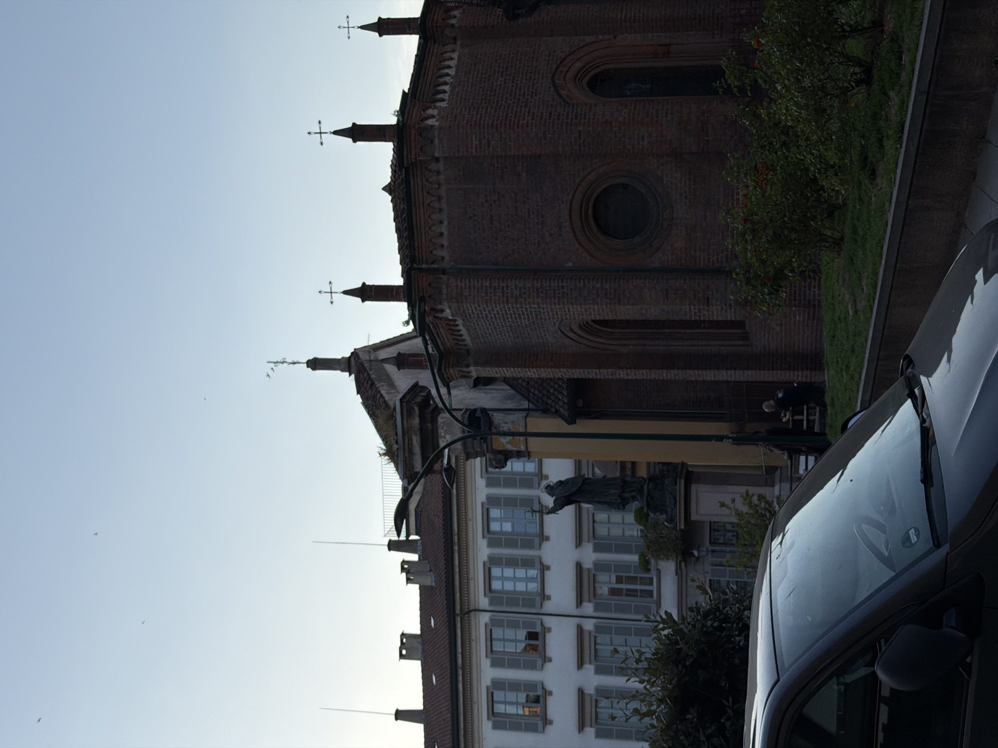 A long shot shows a tall brick building, likely a church or cathedral, situated next to a lighter-colored building with many windows, possibly an apartment complex. The tops of both buildings are visible against a pale sky. The foreground contains a portion of a gray car. A person is visible near the center of the shot. The location may be Milan, Italy.