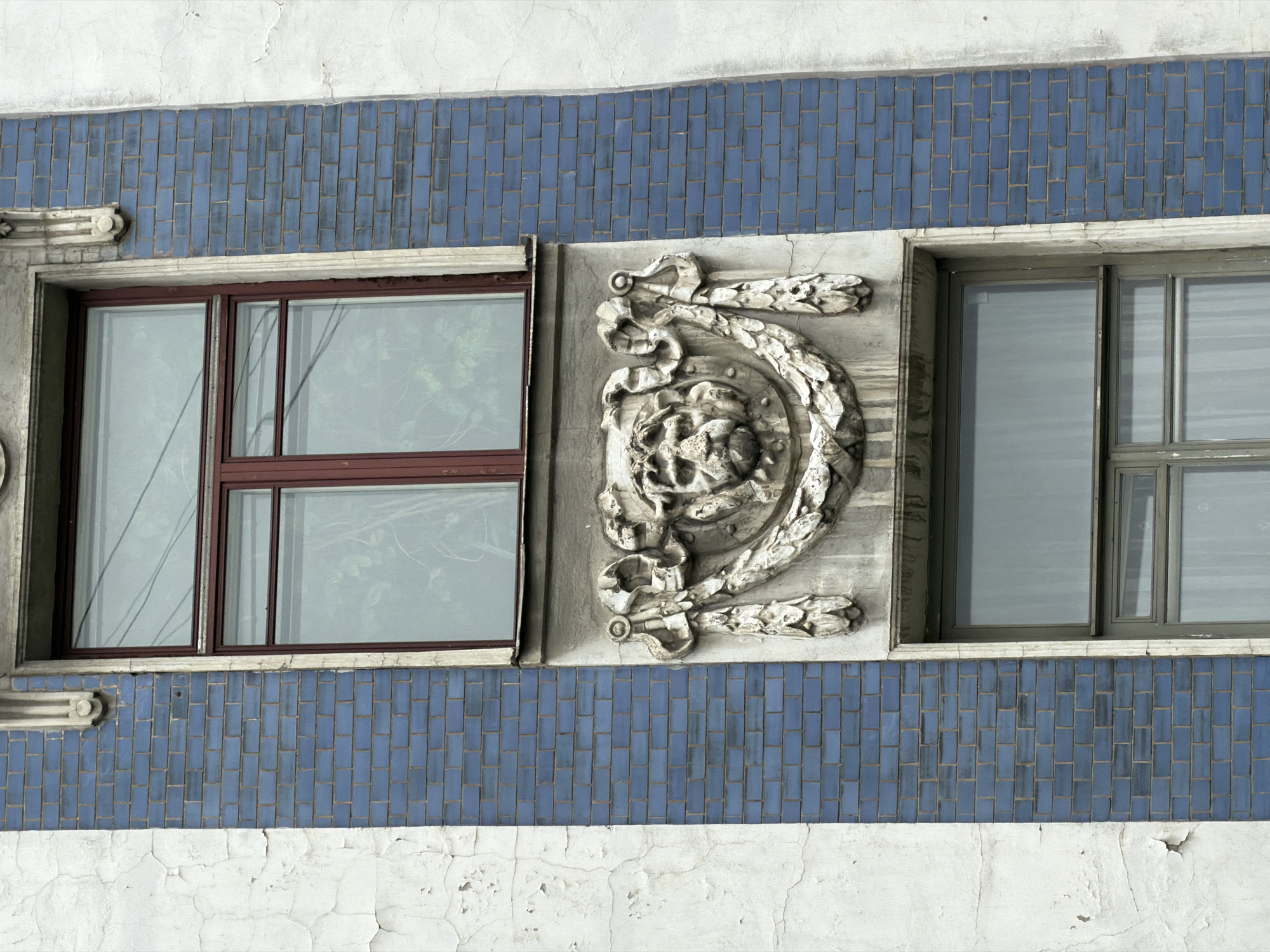 A close-up shot of a building facade in Saint Petersburg, Russia, reveals two windows flanking an ornate, sculpted decorative element. The windows, each framed in a different color (red-brown and grey), reflect the outside world. The wall beneath the windows is coated with a layer of cracked, peeling white paint. Above this, a band of blue rectangular tiles runs horizontally, providing a stark contrast to the crumbling paint below. The sculpted piece between the windows is an elaborate, aged decoration, possibly made of stone or plaster, depicting floral and foliate patterns. It is positioned directly between the two windows, seemingly integrated into the architectural design of the facade. The overall impression is one of aging grandeur, showcasing a blend of architectural features and weathering.