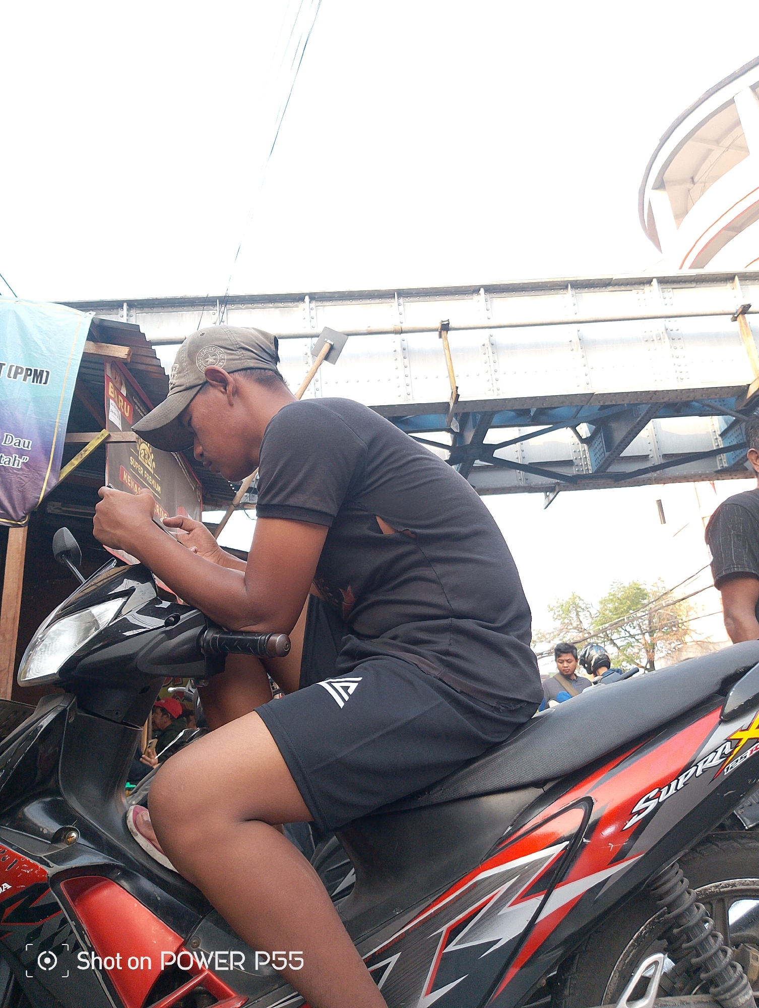 In Surabaya, Indonesia, a person wearing a dark t-shirt and a cap sits on a black and red motorcycle. They are looking down at a mobile device in their hands. The motorcycle has "Supra X" branding on its side. In the background, an elevated railway track structure is visible against a bright sky. Other individuals and trees can be seen in the distance. The scene appears to be a street or roadside.