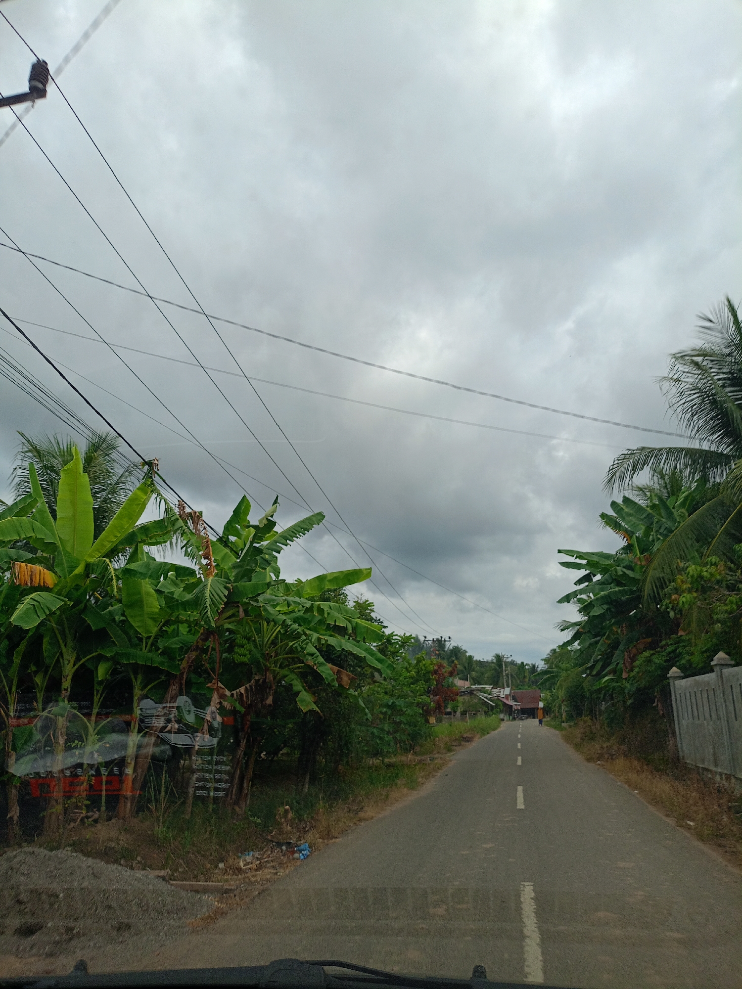 The image shows a road in Seuneuboklhong, Indonesia. The road is asphalt and has a white center line. On either side of the road are banana trees and other vegetation. There are power lines overhead. The sky is cloudy and overcast. A person is walking in the distance down the road.