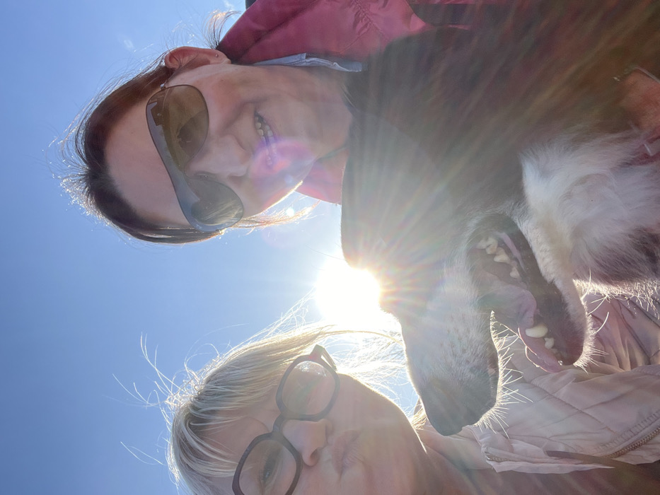 An outdoor, low-angle selfie captured in the city of Chambles, France, features two adult females and one dog against a clear blue sky. The scene is heavily backlit by the sun, producing significant lens flare and a bright halo effect behind the subjects.
The female on the left has blonde hair, wears dark-framed glasses, and a light-colored quilted jacket, posing with pursed lips. The female on the right has dark hair, wears dark aviator-style sunglasses, and a pink jacket over a blue collared shirt, smiling towards the camera.
A black and white dog, appearing to be a medium-to-large breed, is positioned centrally below the women. Its mouth is open, revealing teeth and a portion of its tongue, indicative of panting or an expressive stance. The dog's head is tilted slightly upwards. The subjects collectively occupy the majority of the frame.
