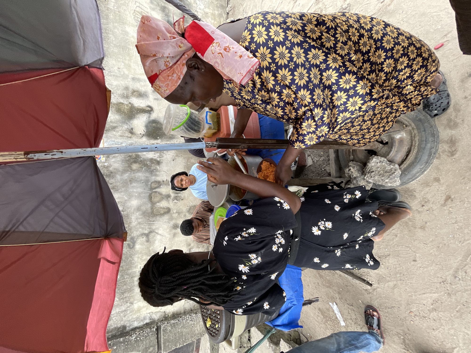 In Ado, Nigeria, two women are engaged in a transaction at an outdoor food stall. The woman on the right, wearing a patterned headscarf and a blue and yellow floral dress, is serving food from a large pot to the woman on the left, who is wearing a black dress with white flowers and is holding a plate of food. Both women are positioned under a large umbrella, which provides shade. In the background, two other individuals, a man and a woman, are visible, seated and observing. The environment appears to be a street scene with a concrete ground and a textured wall behind the stall. The food stall itself features various containers of food and utensils, suggesting a bustling food preparation and sales area.