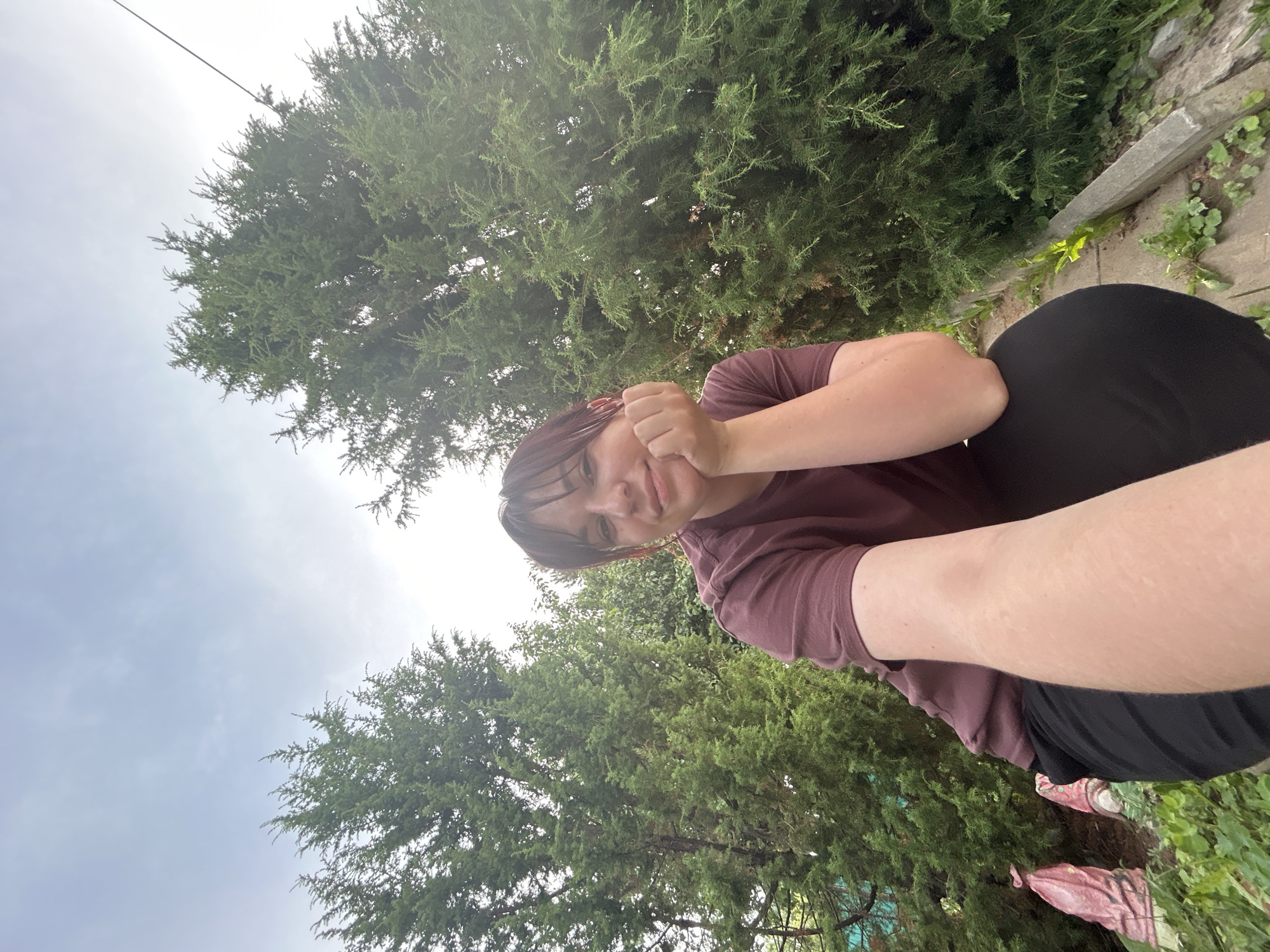 A young person with dark hair is resting their head on their hand, looking directly at the camera. They are wearing a maroon t-shirt and black pants. The person is positioned outdoors, with a large green tree filling the background. The sky is visible above the tree, appearing overcast. The lighting suggests it is daytime, likely late afternoon or early evening. The scene appears to be a yard or garden area, with some stone edging and greenery visible. The person's expression is relaxed and somewhat contemplative. No visible text is present in the image.