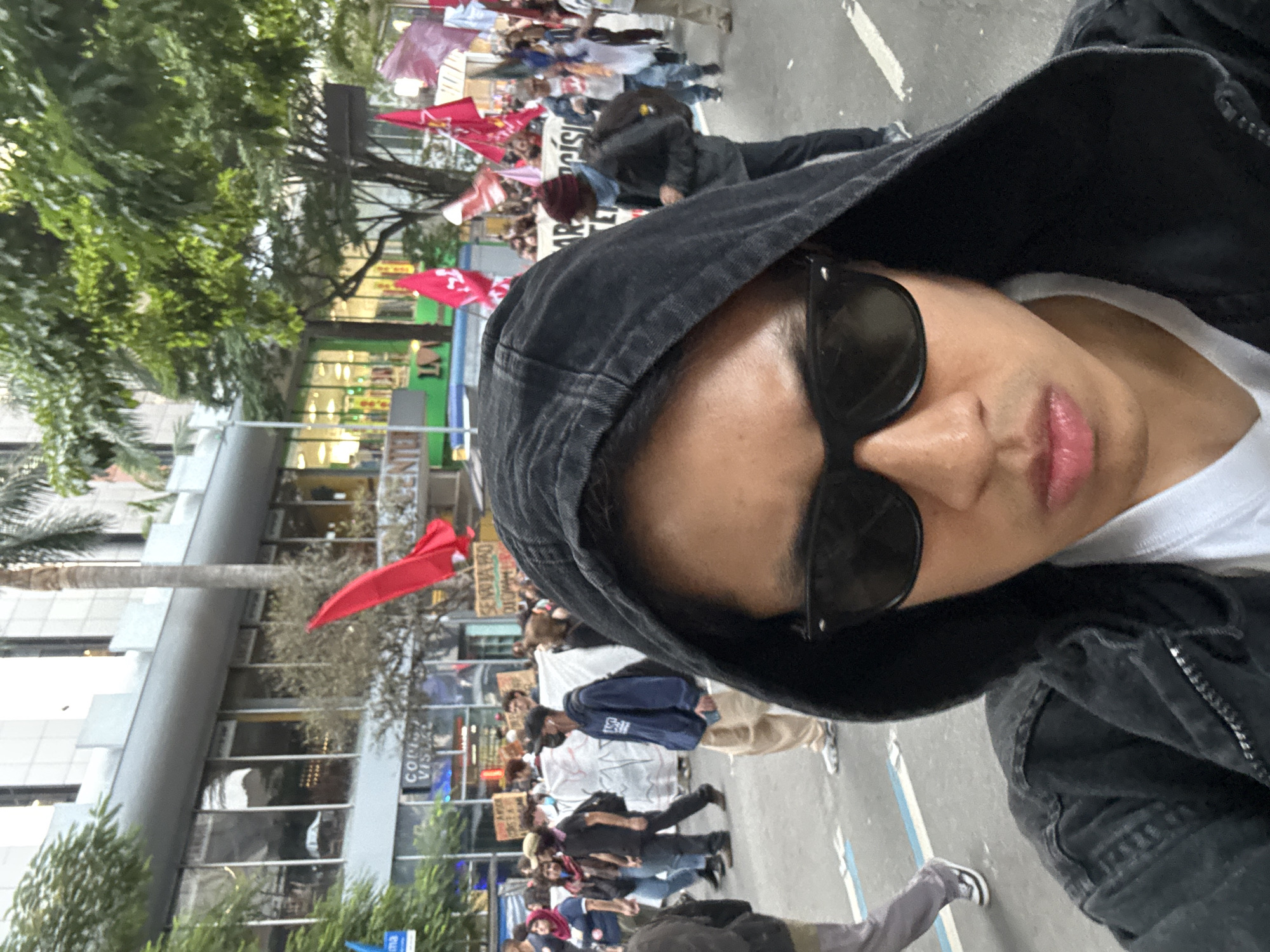 A selfie taken during a protest in São Paulo, Brazil. The foreground features a person wearing sunglasses and a black hoodie, suggesting a potentially incognito presence at the event. A crowd is visible in the background, many holding red flags, indicating a political demonstration. Banners with text are visible among the protestors. Buildings and trees can be seen in the background, indicating an urban environment. The angle and composition of the photo suggest the person is an active participant in the event.