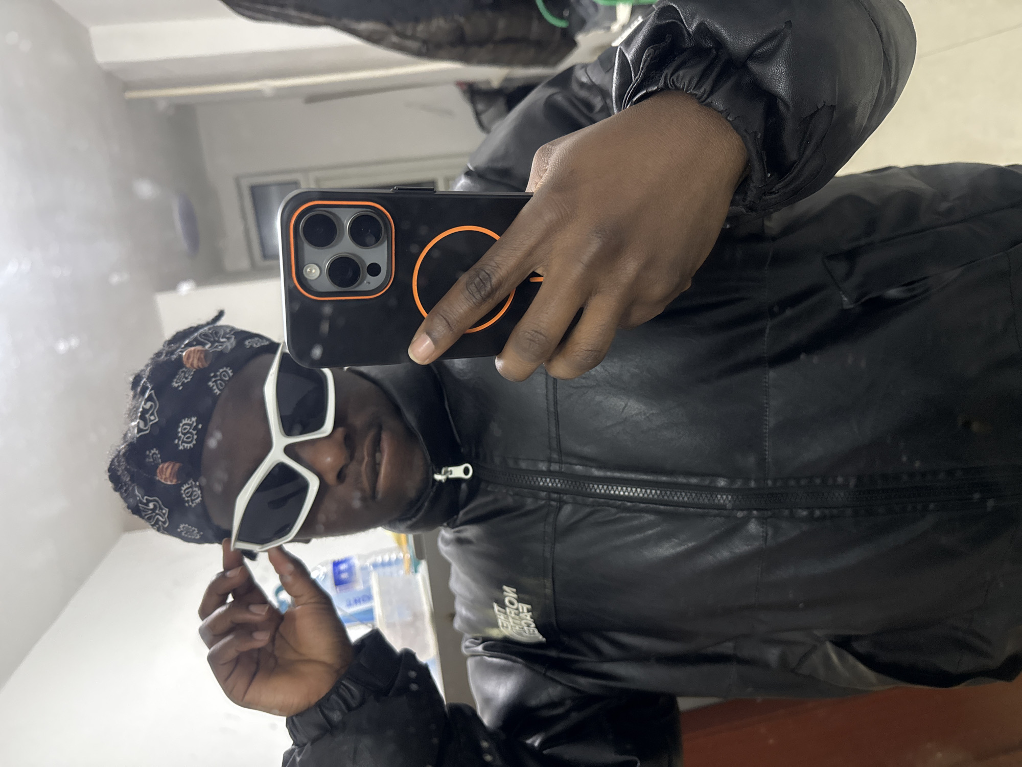 A man is taking a selfie in a mirror. He is wearing a black leather jacket, a bandana, and white sunglasses. He is holding a smartphone with a black case that has an orange ring on the back. The background shows a wall, and a shelf with some items. The location cannot be determined from this image.