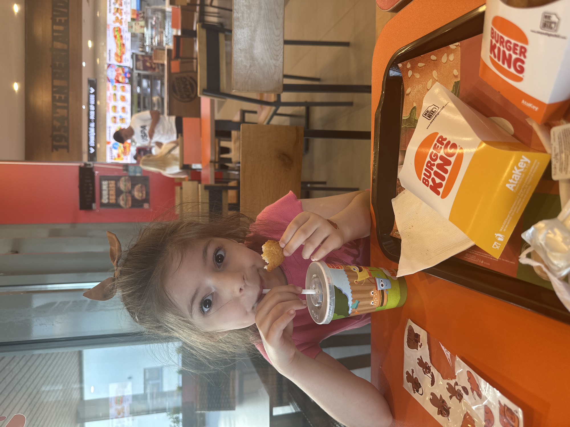 A child is seated at an orange table at a Burger King restaurant in Manisa, Turkey. The child has light brown hair with a bow and is wearing a pink shirt. They are holding a chicken nugget in their left hand and drinking from a decorated cup with a straw in their right hand. On the table is a tray with Burger King food containers, including a box of nuggets, a box of fries, and a cup. Sticker sheets featuring cartoon animals are also on the table. In the background, other tables and chairs are visible, along with a Burger King ordering area. The restaurant's slogan "1954 TEN BERIJALEVD" is displayed on the wall.