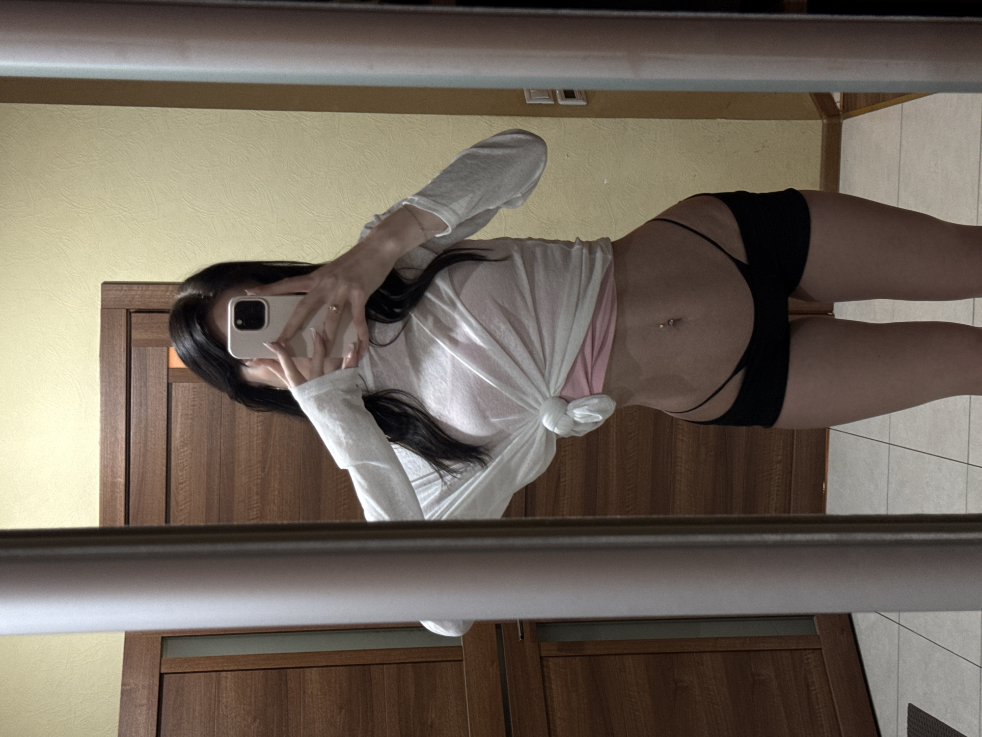 A young woman with long, dark hair is taking a mirror selfie in a room. She is wearing a white, sheer long-sleeved top tied at the waist over a pink undergarment, and black shorts. She is holding a smartphone in her right hand, covering her face. A ring is visible on her left hand. The room has wood-paneled walls and what appears to be a closet door to the left. The floor is tiled. The image was taken in the city of Khimki, Russia.