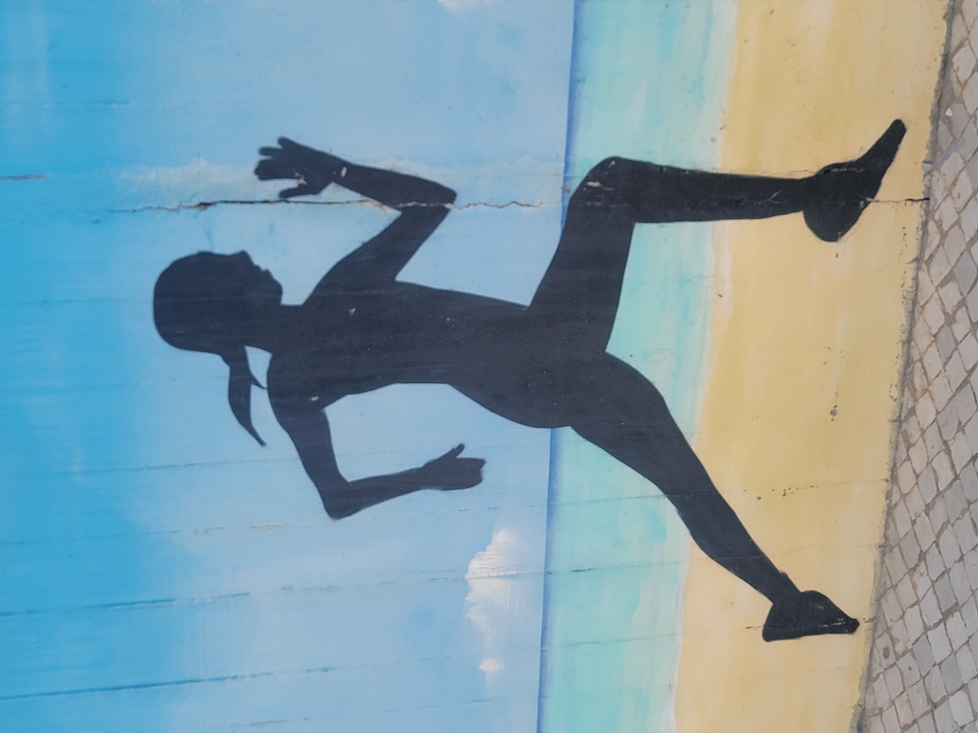 The media file shows a mural painting on a wall. The mural depicts a silhouetted figure of a woman running. She is depicted in motion, with her arms and legs extended as if mid-stride. The background of the mural features a sky with clouds on the left, painted in shades of blue, and a beach with sand on the right, painted in yellow and light brown. The ground beside the beach is a mosaic of light gray and white tiles. The mural appears to be outdoors, possibly on a promenade or wall facing the sea in Quarteira, Portugal, given the beach and tiled ground. The running figure suggests an activity or event related to fitness, leisure, or sports. There is no visible text in the image. The overall impression is one of movement and a coastal setting.