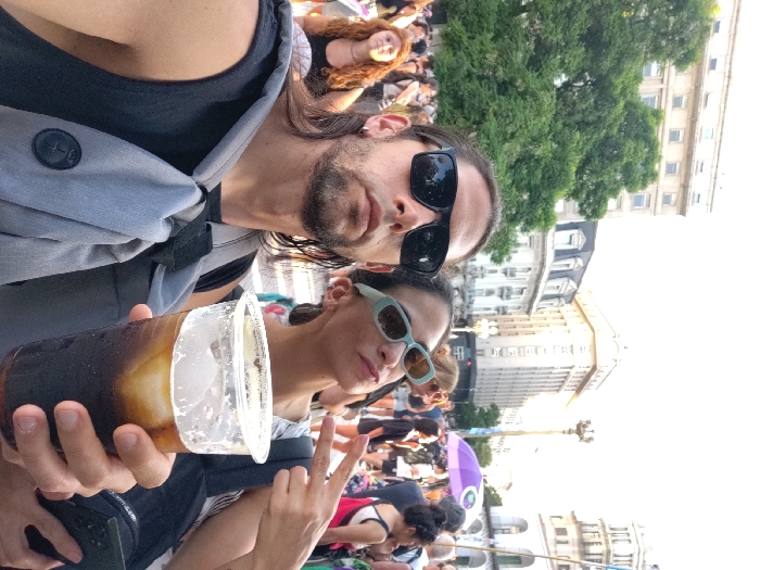 A man and a woman pose for a selfie in a crowded outdoor setting. The man wears sunglasses and a gray bag, while holding a large drink with ice. The woman is wearing pale blue sunglasses and flashes a peace sign. A large crowd of people is visible in the background, along with trees and buildings, possibly in a city square. The image is slightly rotated.