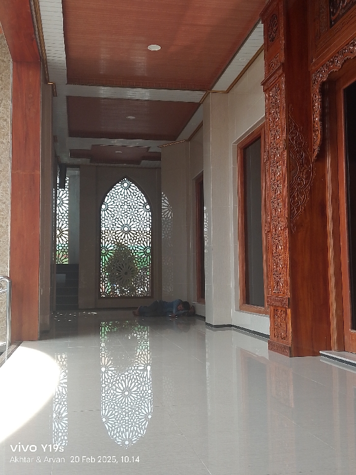 A brightly lit, serene interior corridor, likely within a mosque in Patrol, Indonesia, is depicted in a vertical shot. The smooth, highly polished, light-colored tiled floor creates strong reflections of the surroundings. In the background, large arched screens or windows feature intricate geometric patterns, reminiscent of traditional Islamic art, and these patterns are clearly reflected on the floor. One individual, dressed in dark clothing, is seen lying on the floor near these patterned screens, appearing to be resting or sleeping.
The corridor features light-colored tiled walls and a multi-layered ceiling with recessed panels finished in a reddish-brown, wood-like material. Recessed lighting is visible in the ceiling. On the right side of the frame, there is a prominent wooden structure with elaborate, deeply carved details, possibly part of a decorative column or a doorway frame. A simple metal handrail is partially visible on the far left.
Bright daylight streams through the patterned screens, casting distinct shadows and vibrant reflections across the reflective floor, indicating a clear and sunny day, likely morning or early afternoon given the angle of light. The bottom left corner of the image contains visible text: "vivo Y19s," "Akhtar & Arvan," and the date and time "20 Feb 2025, 10.14." The overall atmosphere of the scene is peaceful and quiet.