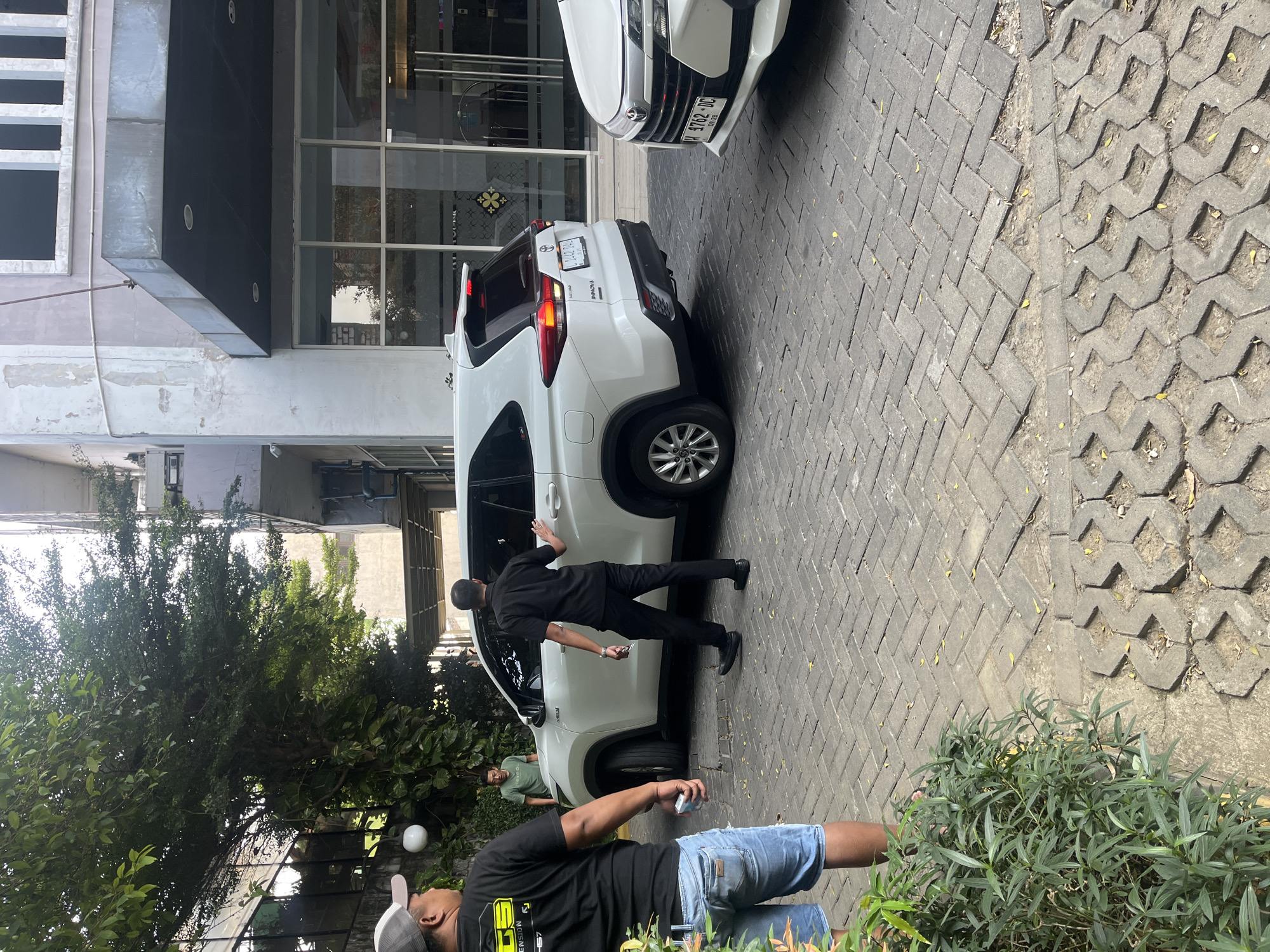 A white SUV, identified as a Toyota Innova, is parked on a brick-paved driveway in front of a building with large glass windows and a dark awning. Two men in black shirts and pants are near the SUV, one appearing to be a driver or valet, with his hand on the rear passenger door. Another man in a black t-shirt and denim shorts is standing to the left, facing away from the camera, holding a pack of cigarettes. A third man in a green t-shirt is standing further back, looking towards the SUV. The scene is outdoors, with trees and foliage visible to the left. The license plate on the Innova reads "L 1442 DLX". A portion of another white car is visible on the right side of the frame. The building's facade shows signs of wear and peeling paint. The ground is paved with interlocking bricks, some of which are arranged in a decorative pattern. The image appears to have been taken in Gapurosukolilo, Indonesia, given the vehicle's license plate format.