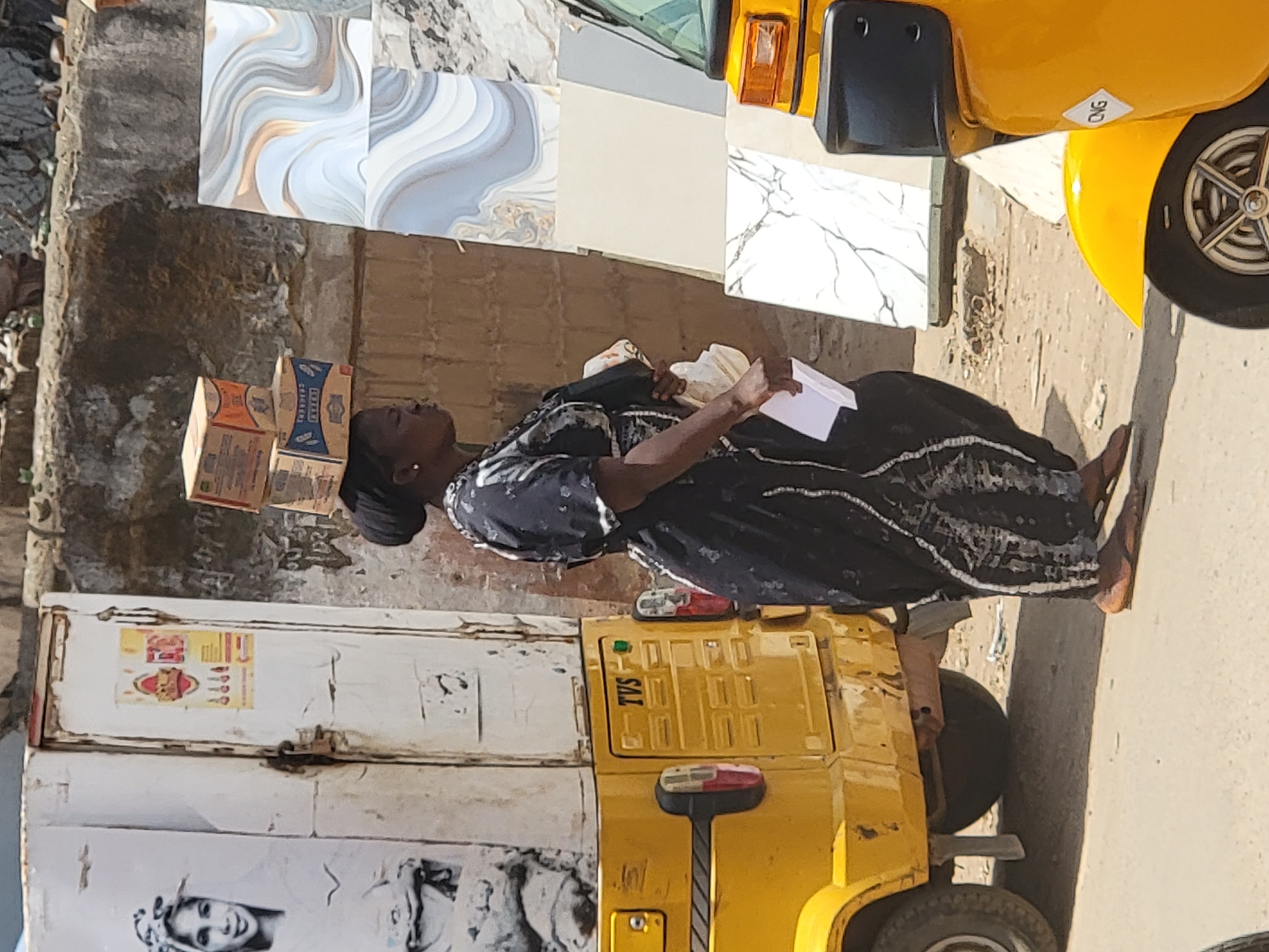 A woman balances three boxes on her head while walking on a street in Maiburiji, Nigeria. She carries a white paper in her left hand. She wears a dark-colored dress with light-colored accents. A yellow auto-rickshaw (TVS brand) is partially visible to her right. To her left is a white panel with advertising posters, and behind her are stacked tiles of various designs. The environment appears to be an urban street.
