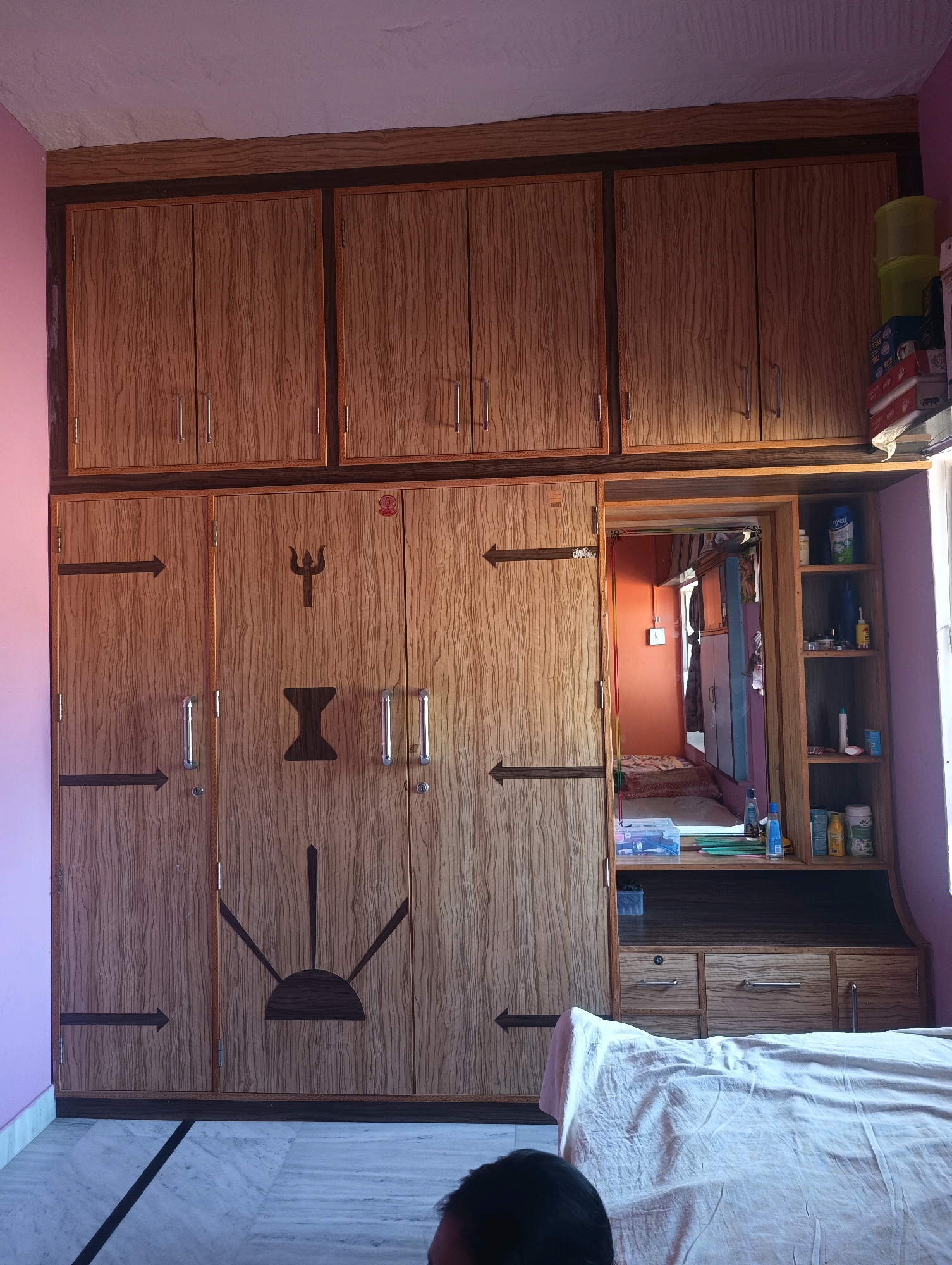 The image shows a large, wooden wardrobe with multiple doors and compartments. The wardrobe is located in a room with light purple walls and a marble floor. The wardrobe is decorated with a trident symbol and other decorative patterns. On the top of the wardrobe, there are several stacks of items, including bottles and boxes. To the right of the wardrobe, there is a smaller cabinet with shelves, displaying various toiletries and containers. A mirror is embedded in the center of the wardrobe, reflecting a room with orange walls and a bed. A portion of a person's head is visible in the lower right corner of the image. The scene appears to be indoors, possibly a bedroom or dressing area, in Chhapri Bari, India.
